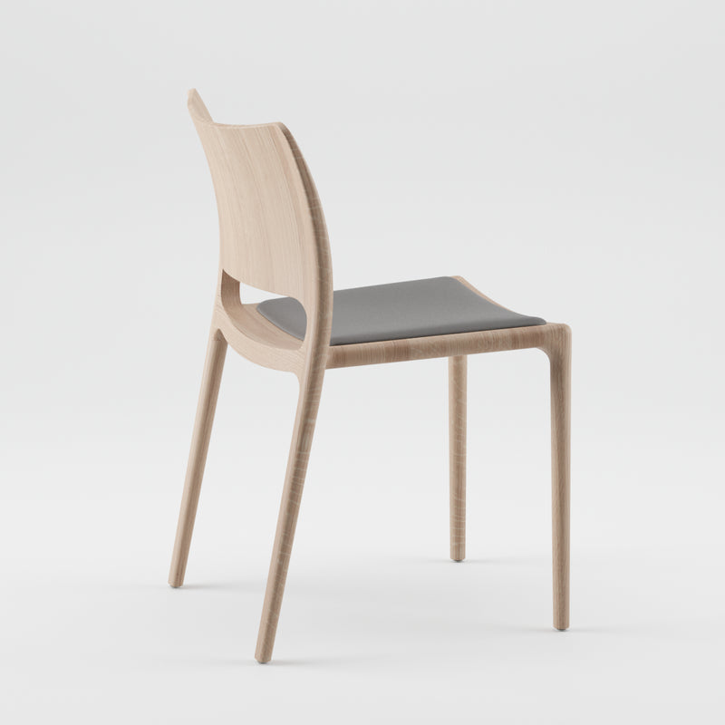 The Artisan LATUS Chair Upholstered is a minimalist design crafted from solid wood, featuring a light wood finish and a simple curved backrest treated with natural oils. It has a smooth, dark gray cushion and is showcased against a plain white background.