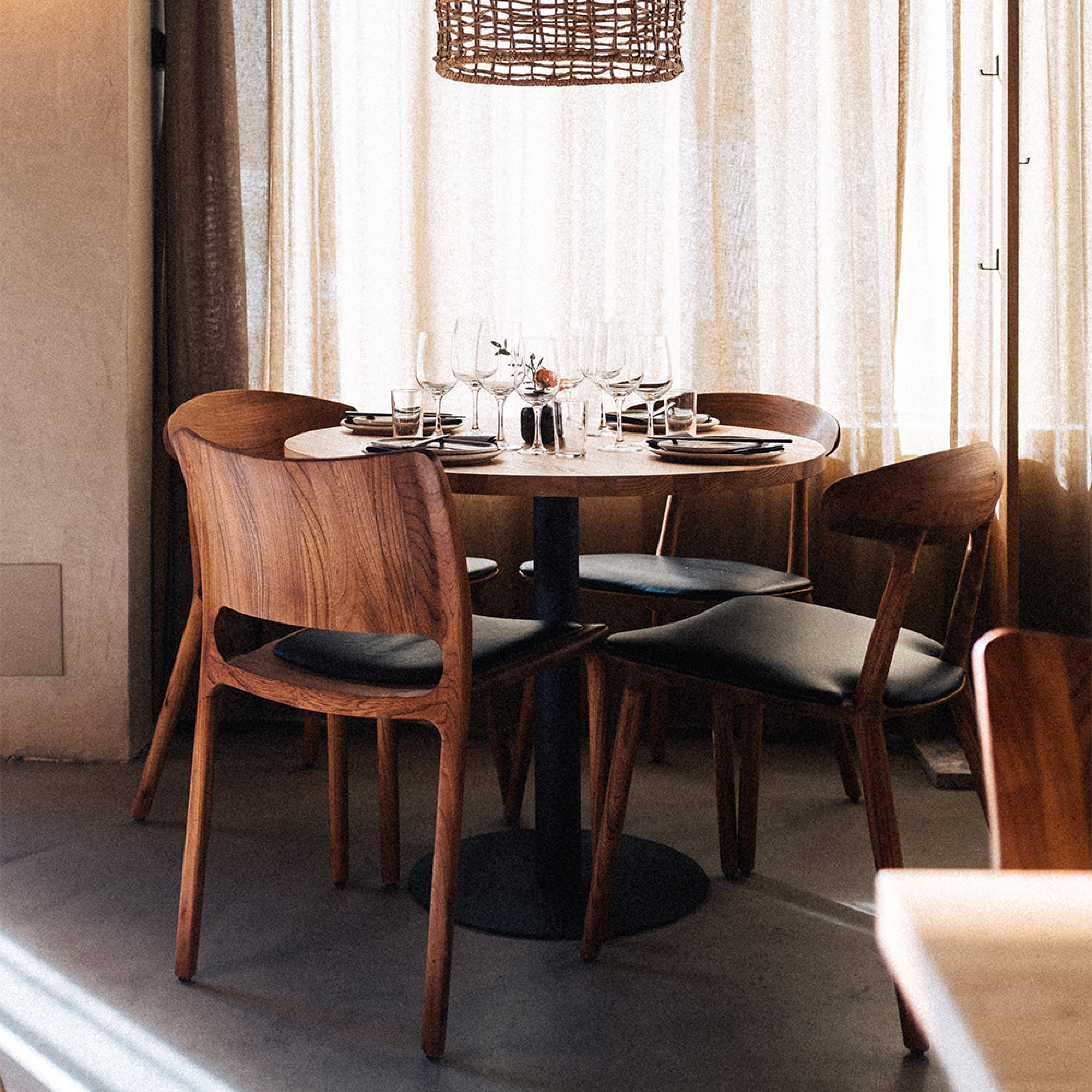 In a warmly lit dining room, the Artisan LATUS Chair Upholstered pairs beautifully with a round solid wood table set for four, adorned with elegant glassware and a small centerpiece. Light streaming through beige curtains adds to the cozy, eco-friendly ambiance.