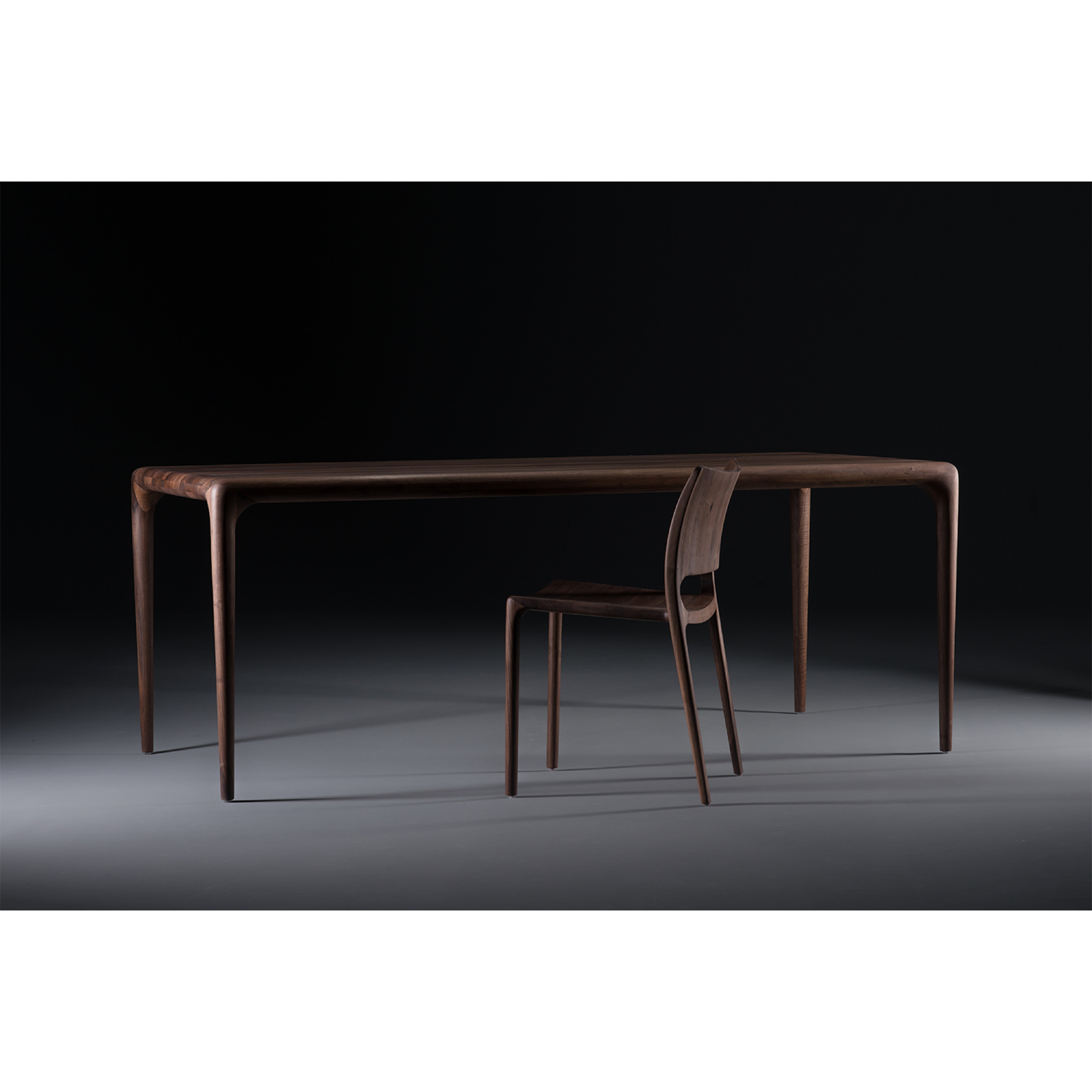 The LATUS Chair by Artisan is a sleek, modern solid wood set with smooth, rounded edges and slender legs. Enhanced with natural oils, the chair tucks partially under the table against a dark, minimalist backdrop.