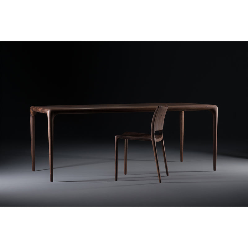 The LATUS Chair by Artisan is a sleek, modern solid wood set with smooth, rounded edges and slender legs. Enhanced with natural oils, the chair tucks partially under the table against a dark, minimalist backdrop.