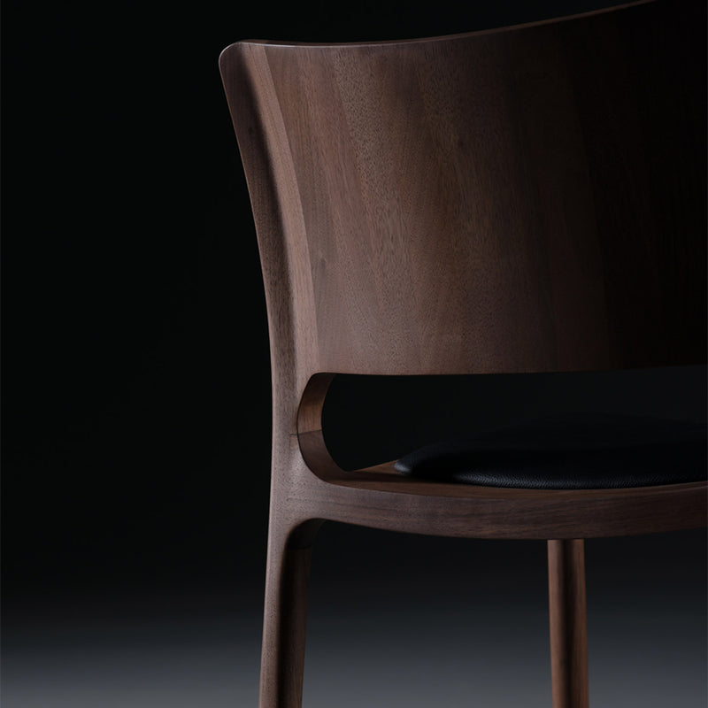 The Artisan LATUS Chair Upholstered features a minimalist design with a curved backrest and a black cushioned seat. Crafted from eco-friendly solid wood and finished with natural oils, its smooth finish stands out against the dark background, highlighting its modern elegance.