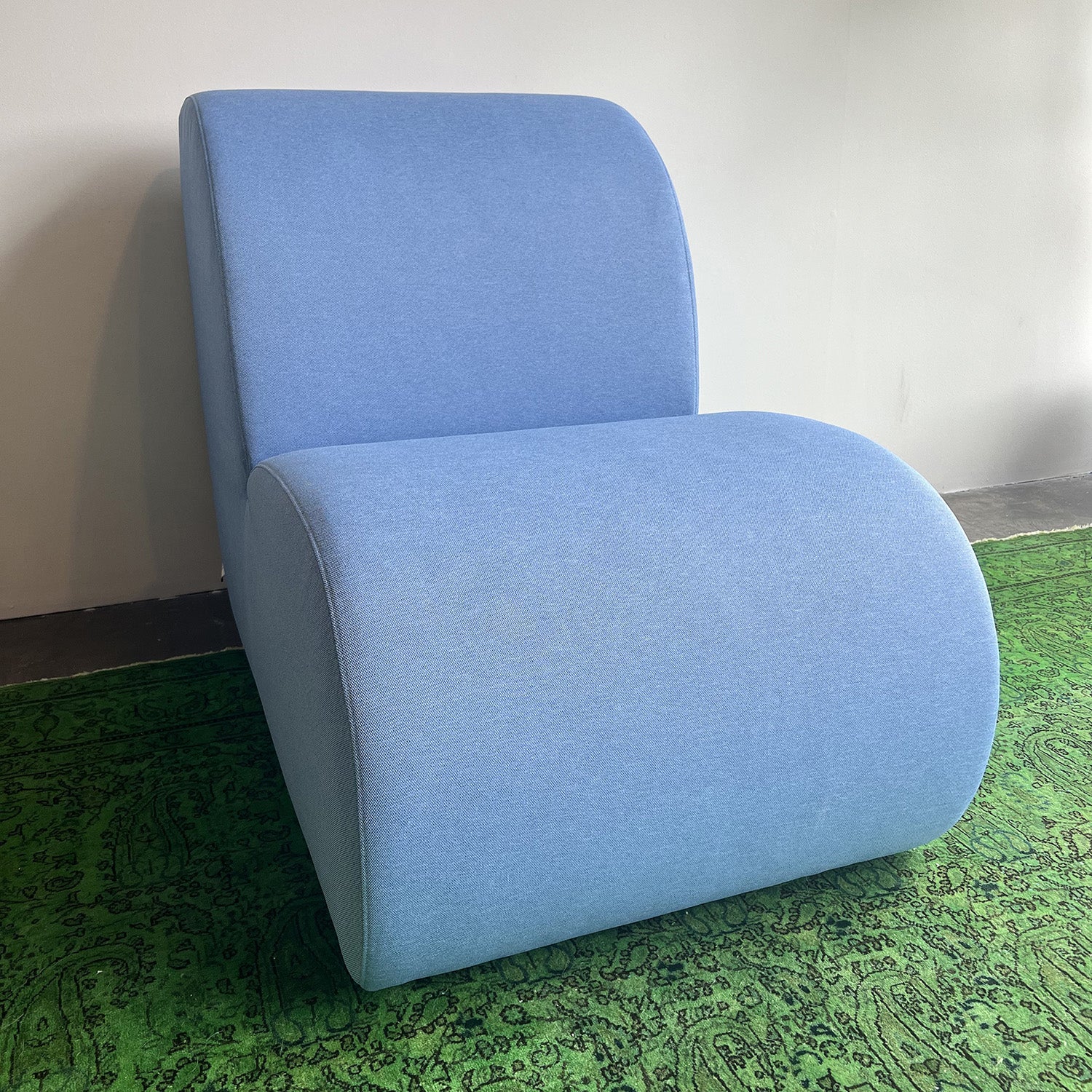 A modern Levi Seating Lounge Chair by Objekte unserer Tage, featuring a curved minimalist design in blue, sits on a green patterned rug. It showcases handcrafted upholstery with an arched backrest and seat, offering a sleek look against the plain light-colored wall.