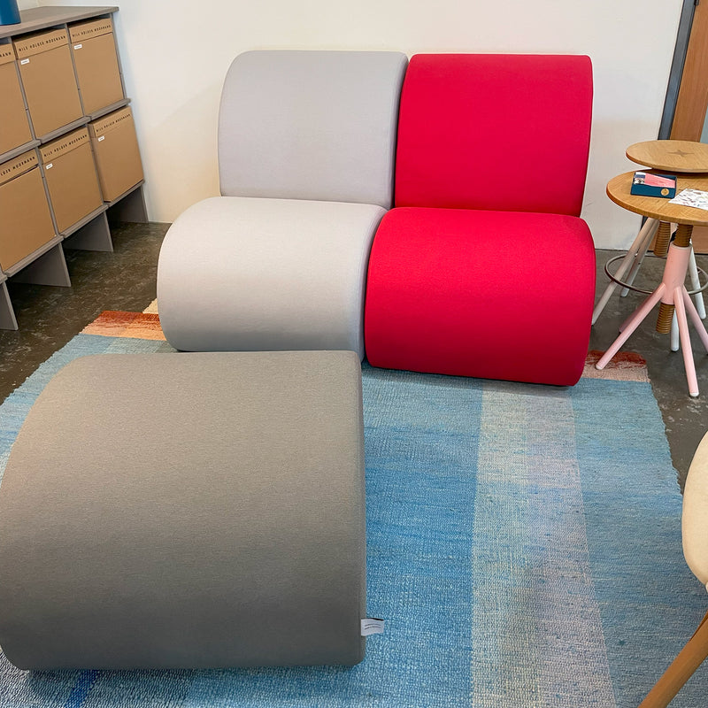 Versatile furniture shines with the Levi Seating Lounge Chair from Objekte unserer Tage. It features curved gray, red, and beige seats on a striped blue and beige rug. A nearby small wooden side table complements part of a storage cabinet.
