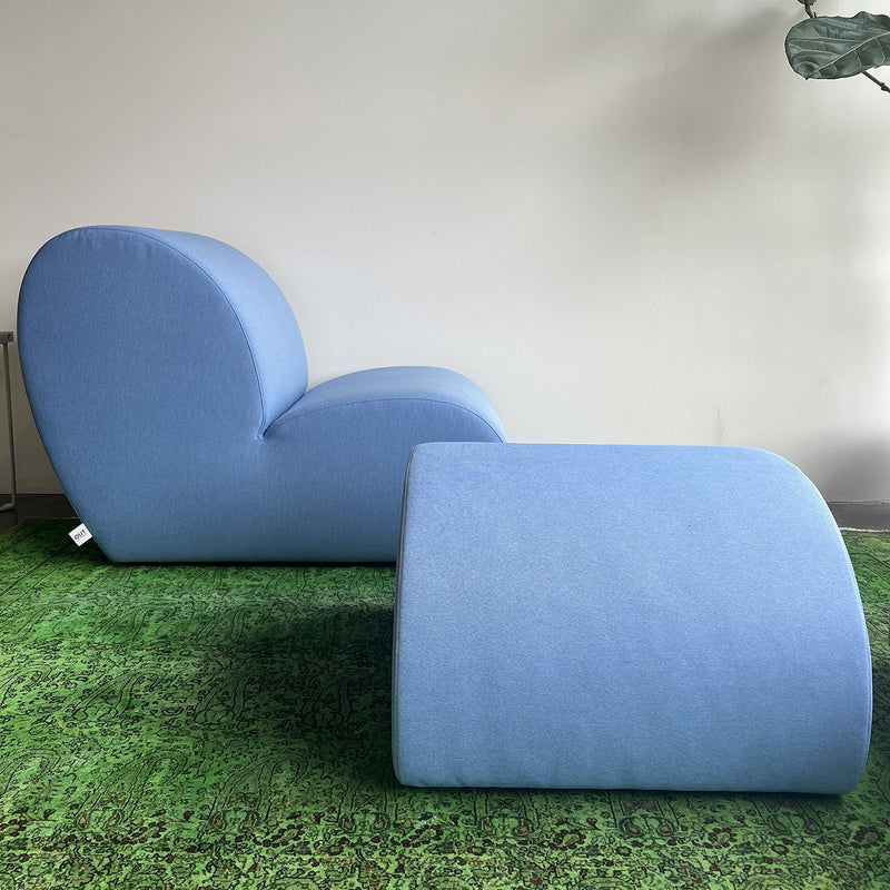 Explore the Levi Seating Lounge Chair by Objekte unserer Tage: a modern blue upholstered chair with a matching ottoman featuring minimalist curves, set on a textured green carpet near the wall with hints of plant leaves in the top right corner. Currently on sale.