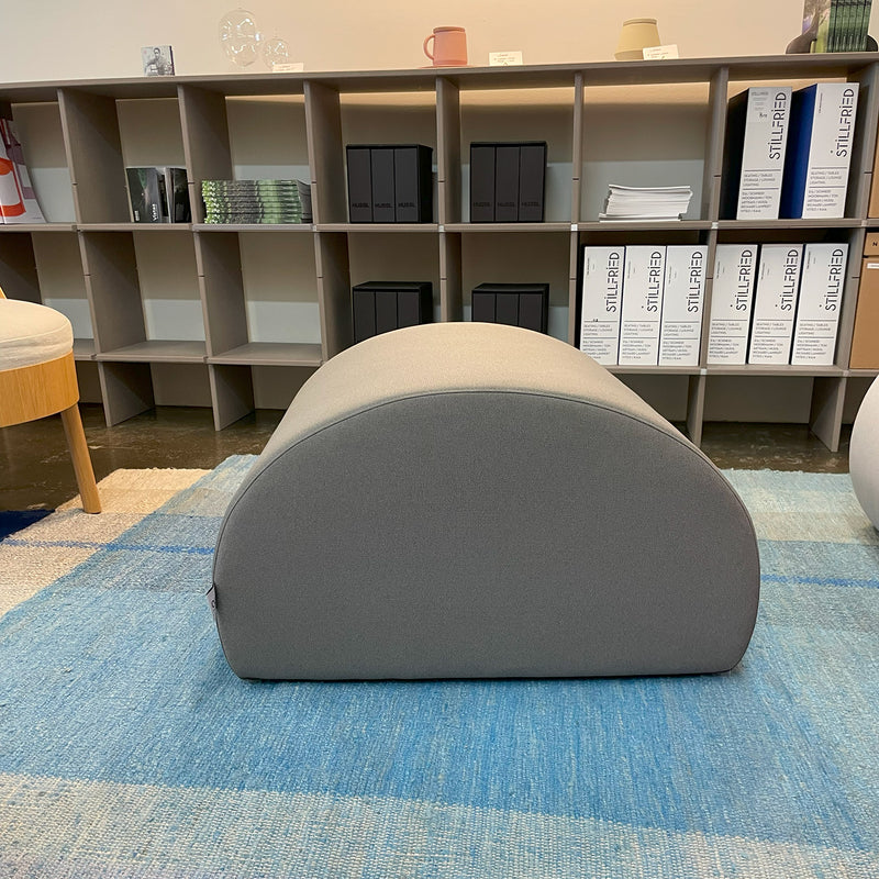 The gray Levi Seating Pouf - SALE by Objekte unserer Tage adds versatility to the room, resting on a blue rug. Behind it, a shelving unit displays files, books, and stilluns labeled white boxes. The minimalist room features beige walls and handcrafted upholstery accents.