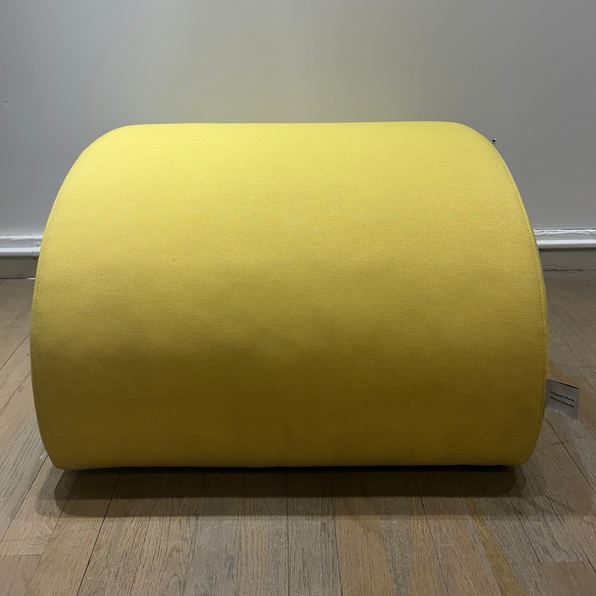 Levi Pouf - Sale, yellow organic-shaped upholstered pouf by Objekte unserer Tage on wooden floor against gray wall.
