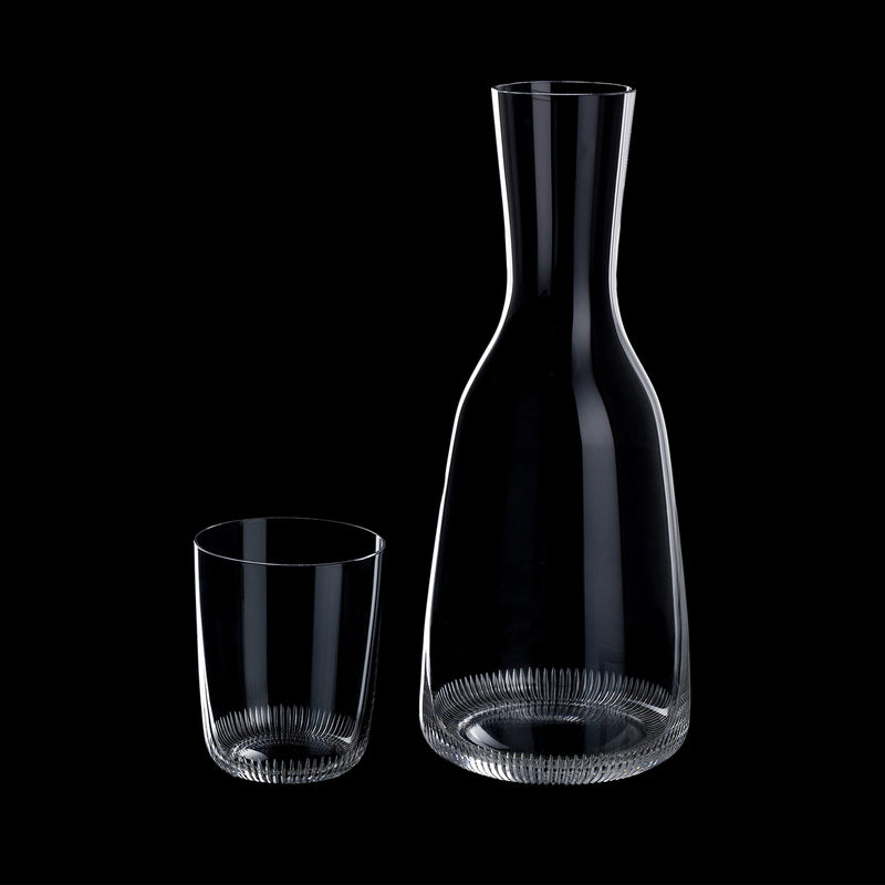 A mouth-blown, lead-free Lobmeyr Drinking Set No. 281 - Grip - Sale, featuring a clear glass carafe and matching tumbler with vertical etched lines, stands empty against a black background. Resembling a whiskey decanter, the taller carafe accentuates their simple elegance and technical aesthetic.