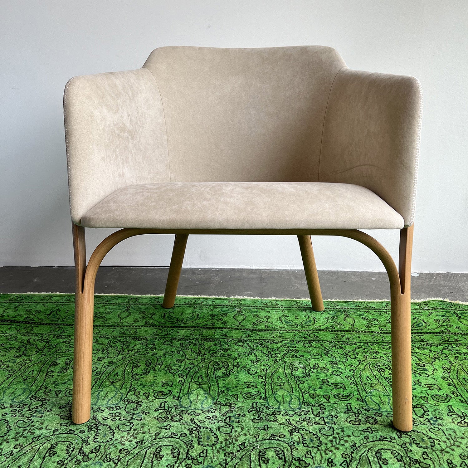 The Lounge Chair Split by TON, featuring comfortable upholstery and manually crafted wooden legs, sits on a vibrant green rug with intricate patterns against a minimalistic light gray wall and dark gray floor.