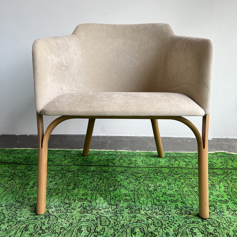 The Lounge Chair Split by TON, featuring comfortable upholstery and manually crafted wooden legs, sits on a vibrant green rug with intricate patterns against a minimalistic light gray wall and dark gray floor.