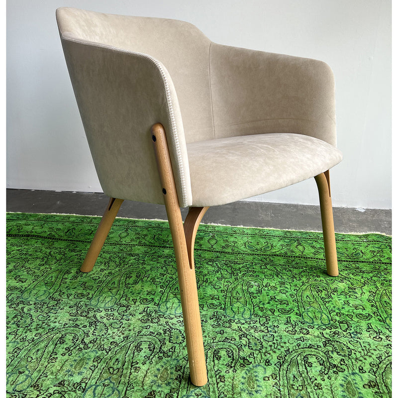 The TON Lounge Chair Split - Sale features sleek, curved lines and cozy beige upholstery with wooden legs. It stands gracefully on a green rug with intricate patterns, creating a simple and modern setting.