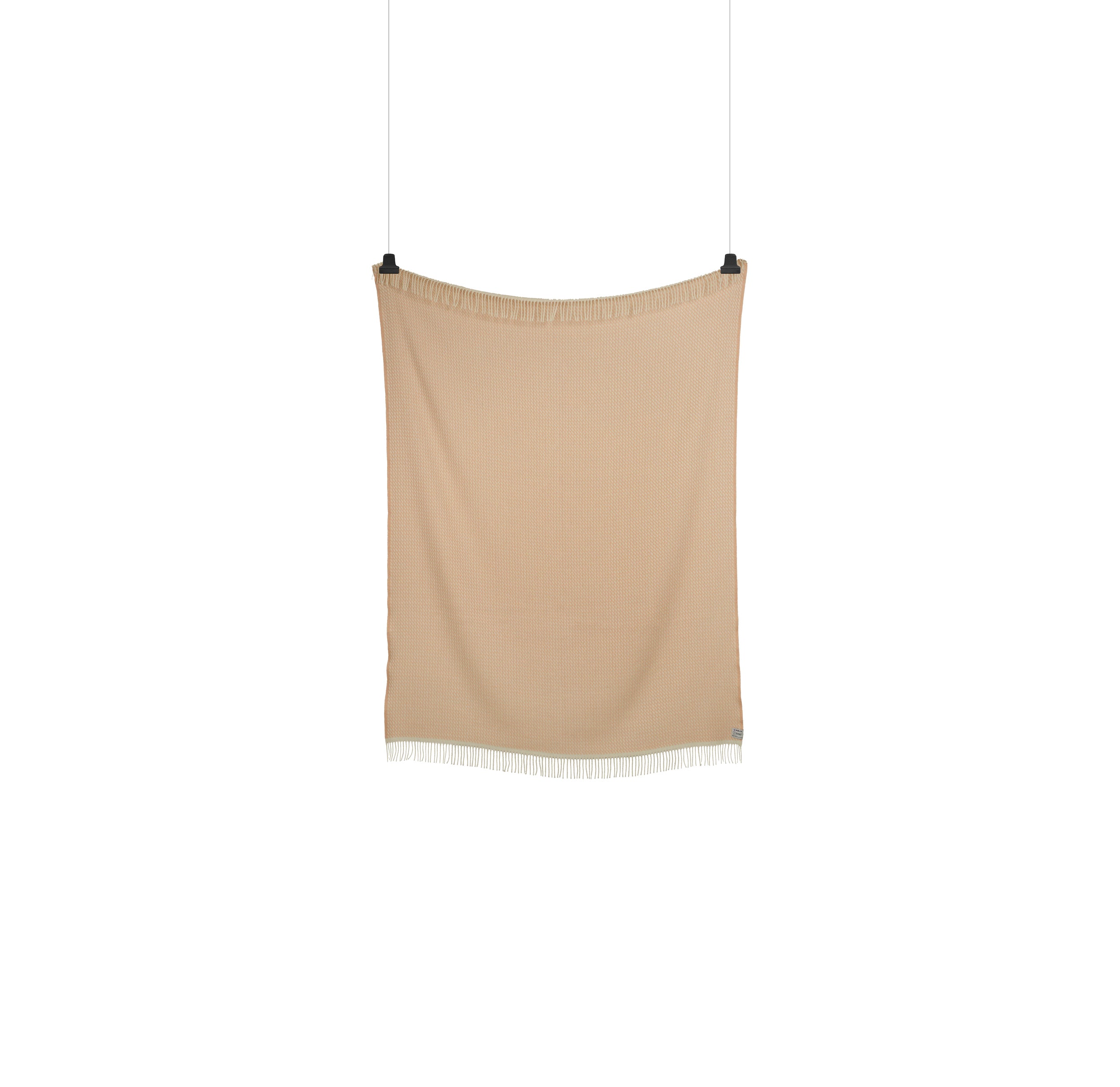 The MELLO Throw by Roros, a beige fabric resembling Norwegian lambswool, hangs against a white backdrop secured by top corner clips and has a fringed bottom edge.