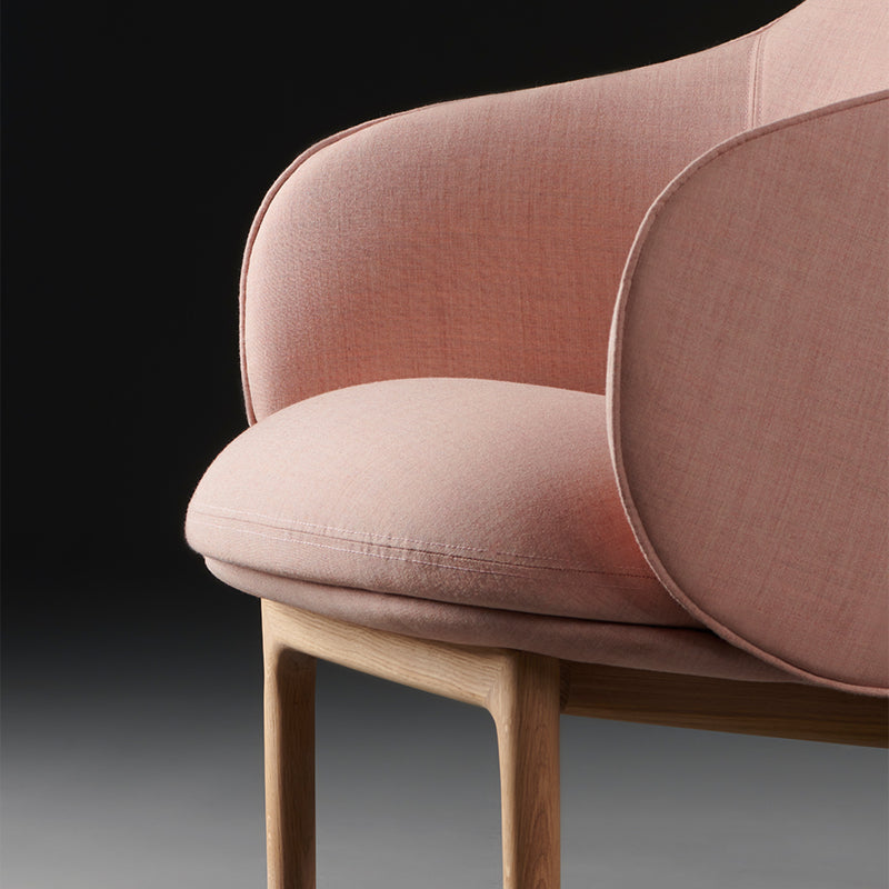 A close-up of the Artisan MELA Lounge Chair High displays a pink upholstered seat with wooden legs set against a dark gray background. Its smooth, rounded cushions and mid-century inspired design emphasize contemporary elegance.