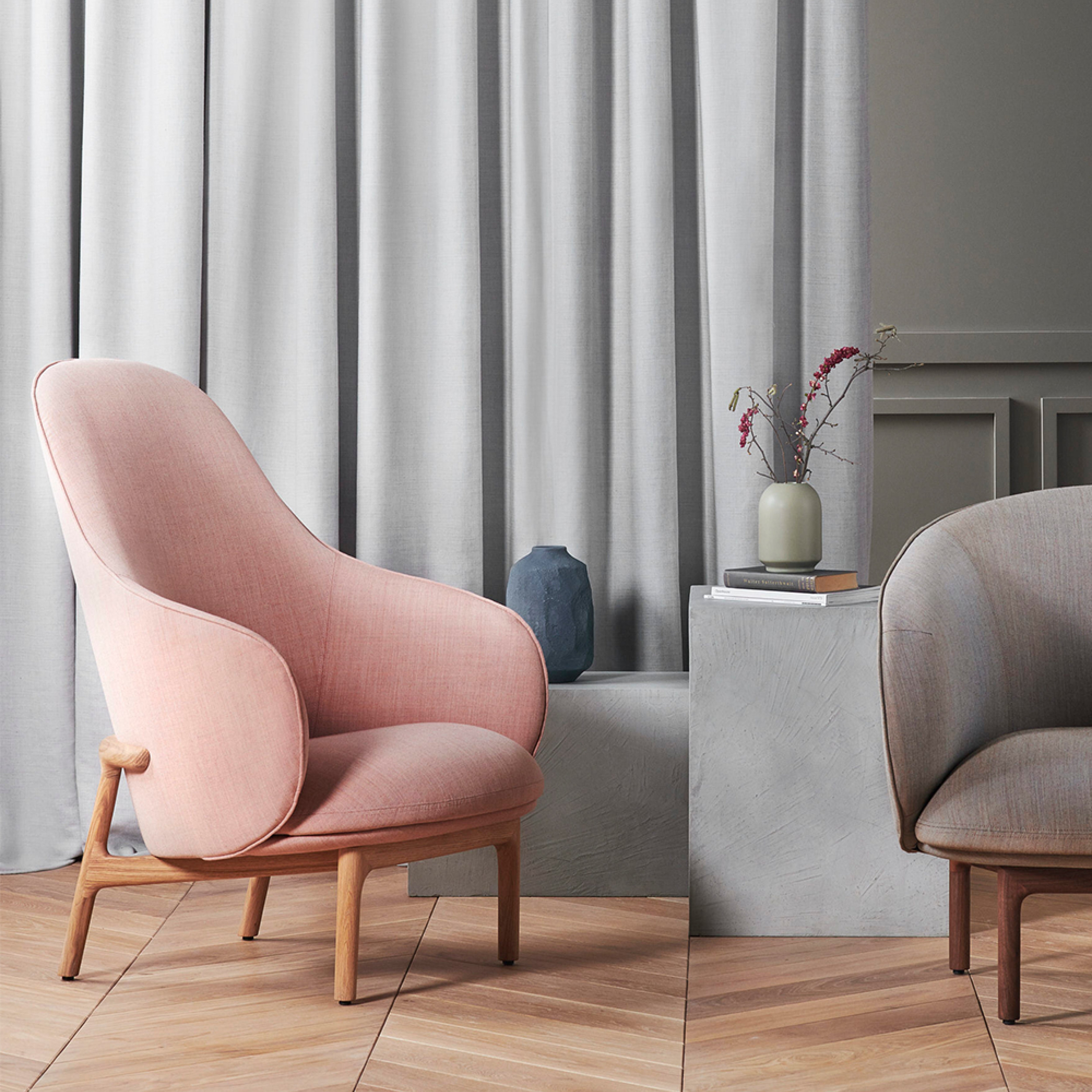In a cozy room, a pink upholstered chair with wooden legs sits on a herringbone wood floor. Partially visible is the MELA Lounge Chair High from Artisan in gray. A white pedestal displays a beige vase with flowers and a dark sculptural vase, while gray curtains hang in the background.