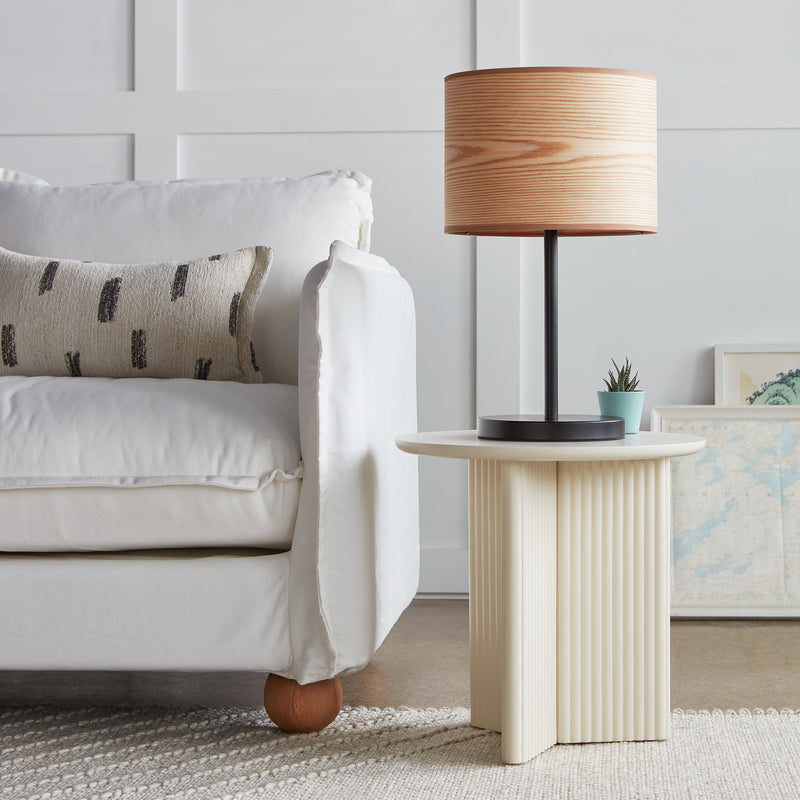 A cozy living room features a white sofa, patterned pillow, round ribbed side table, Gus Modern Milton Table Lamp in wood veneer for warmth, a small potted plant, and framed artwork resting against the wall.