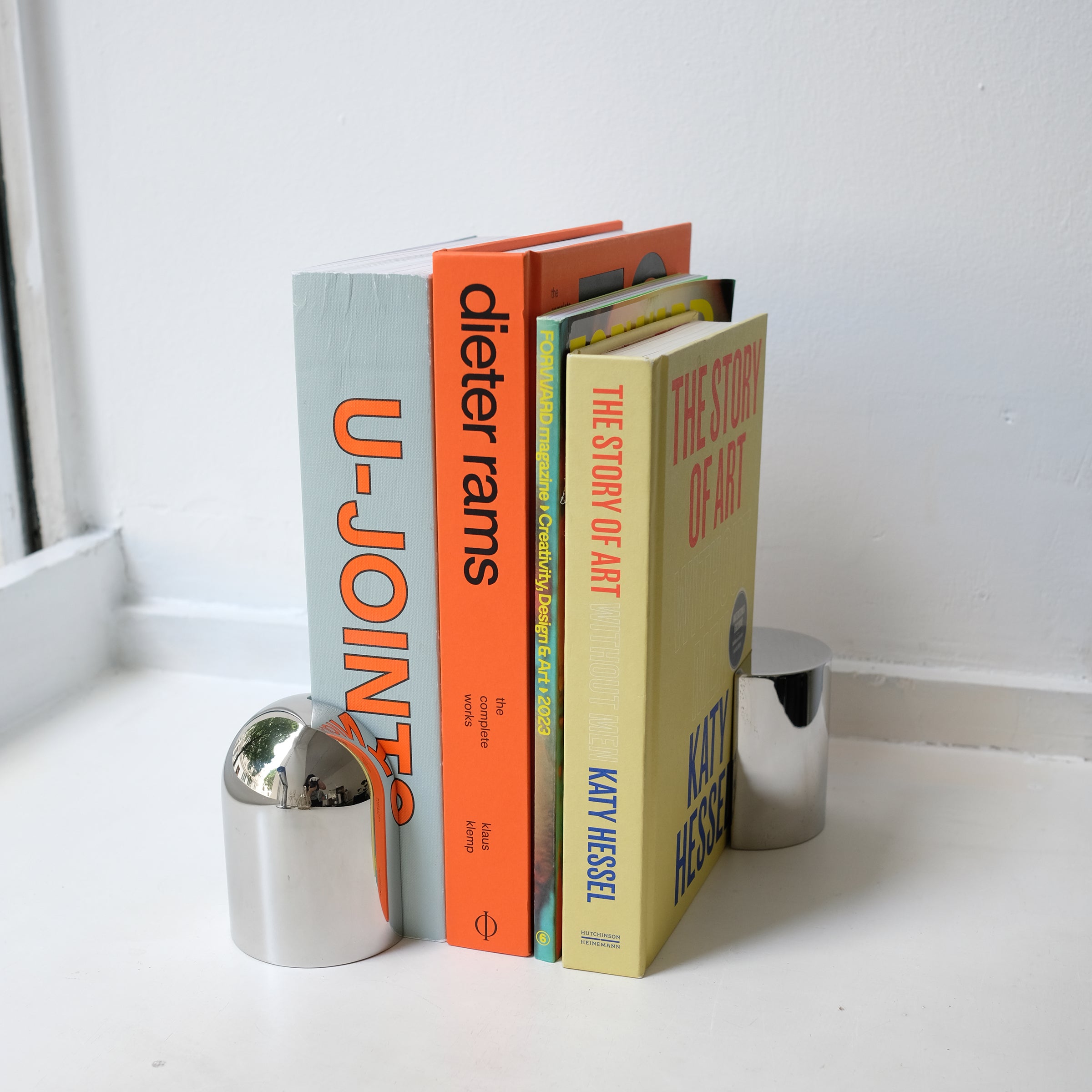 NOOK Bookend B in polished steel holding upright a row of colorful books on a white surface by a wall.