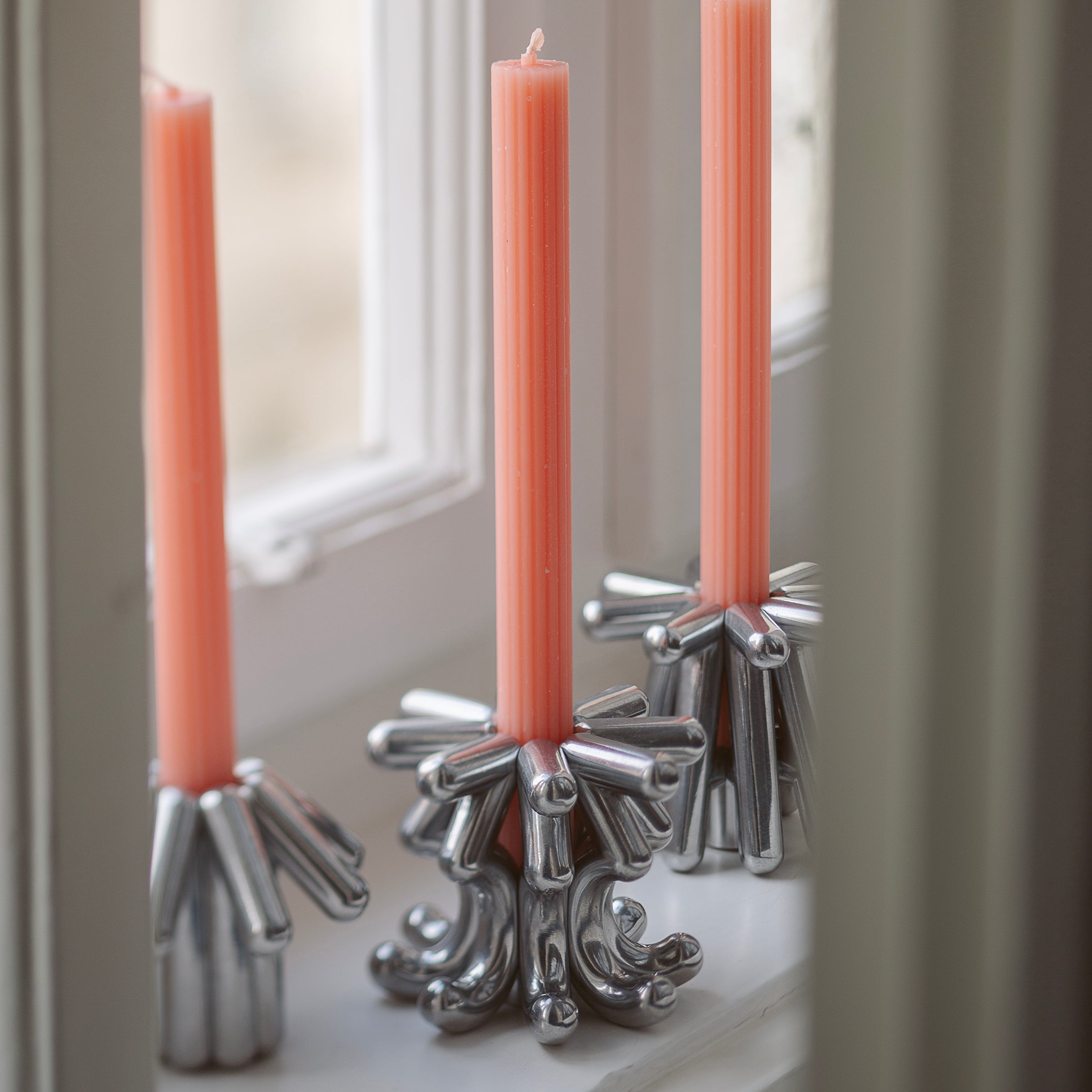 DIGIT FIVE Candlestick in solid aluminum holding a tall, slender pink candle on a windowsill.