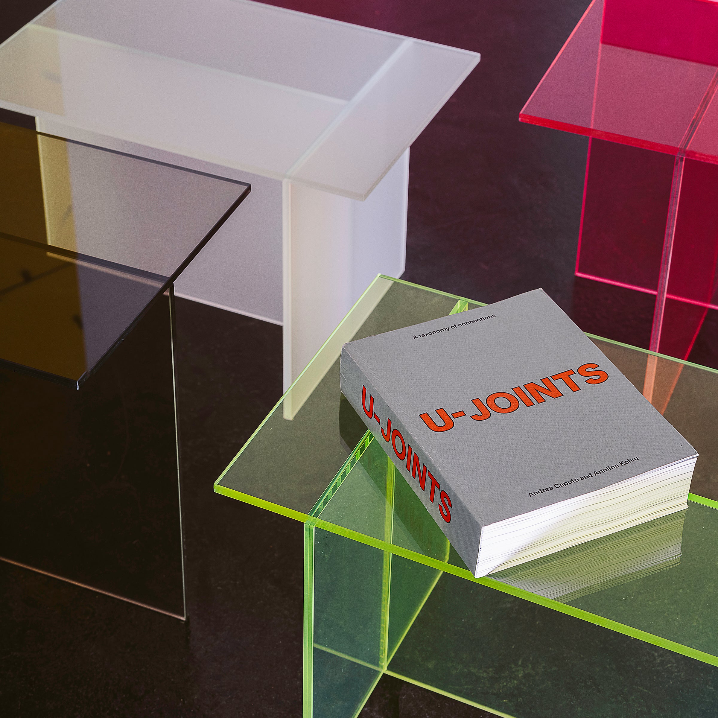 THEO Glass Side Table in multiple colors, featuring intersecting glass elements forming a T shape, shown with a book on top.