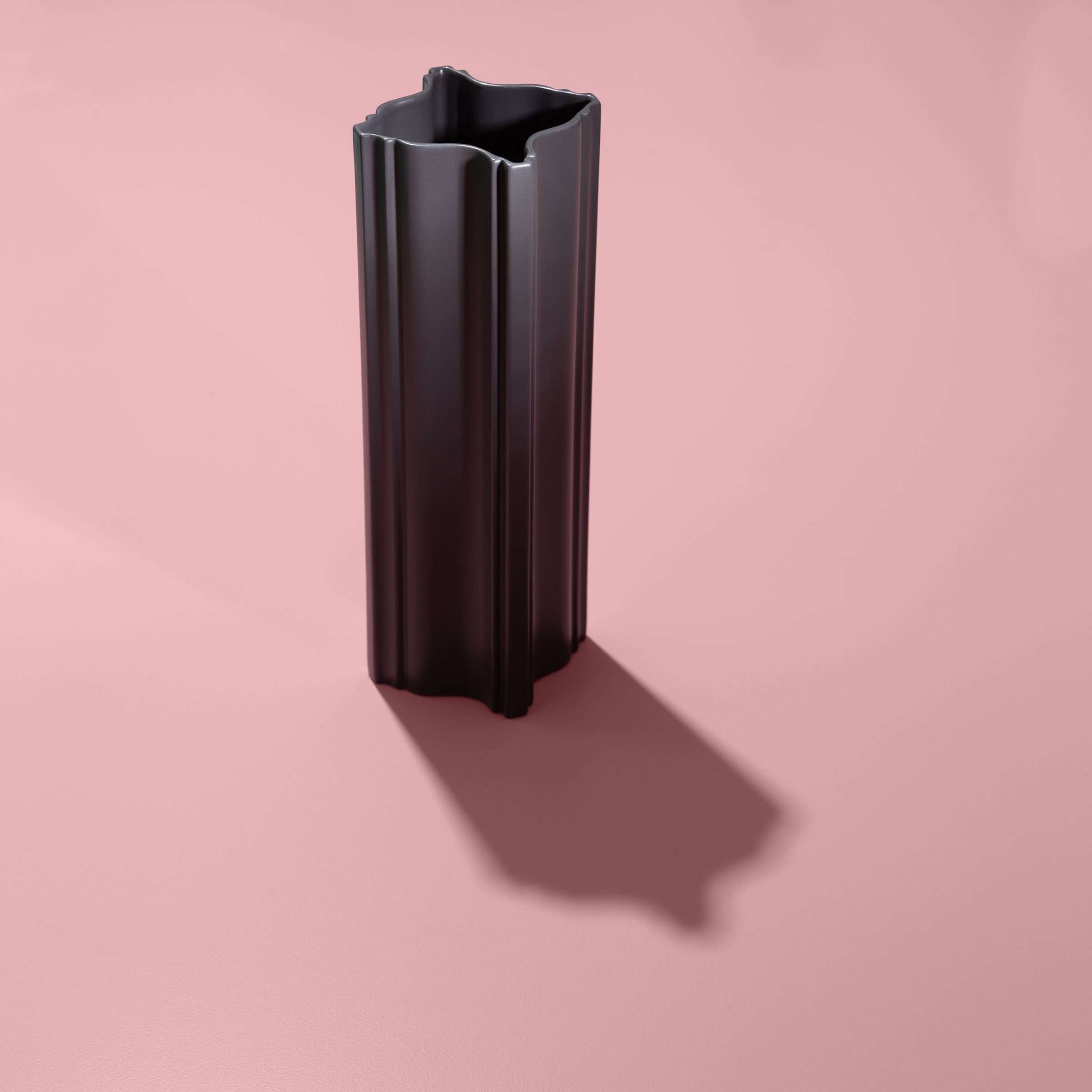 STUCCO Vase in dark grey matte ceramic with architectural, sculptural design on pink background.