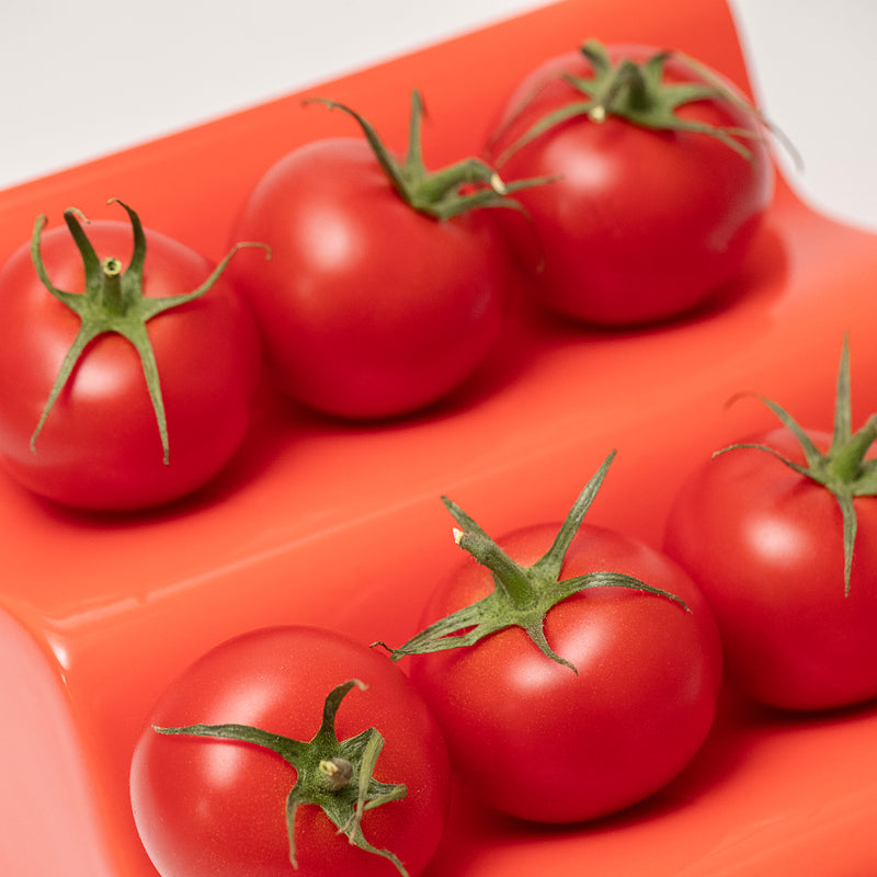 Close-up of red glossy ceramic CASCADE Fruitstand holding fresh ripe tomatoes in a row on a bright surface.