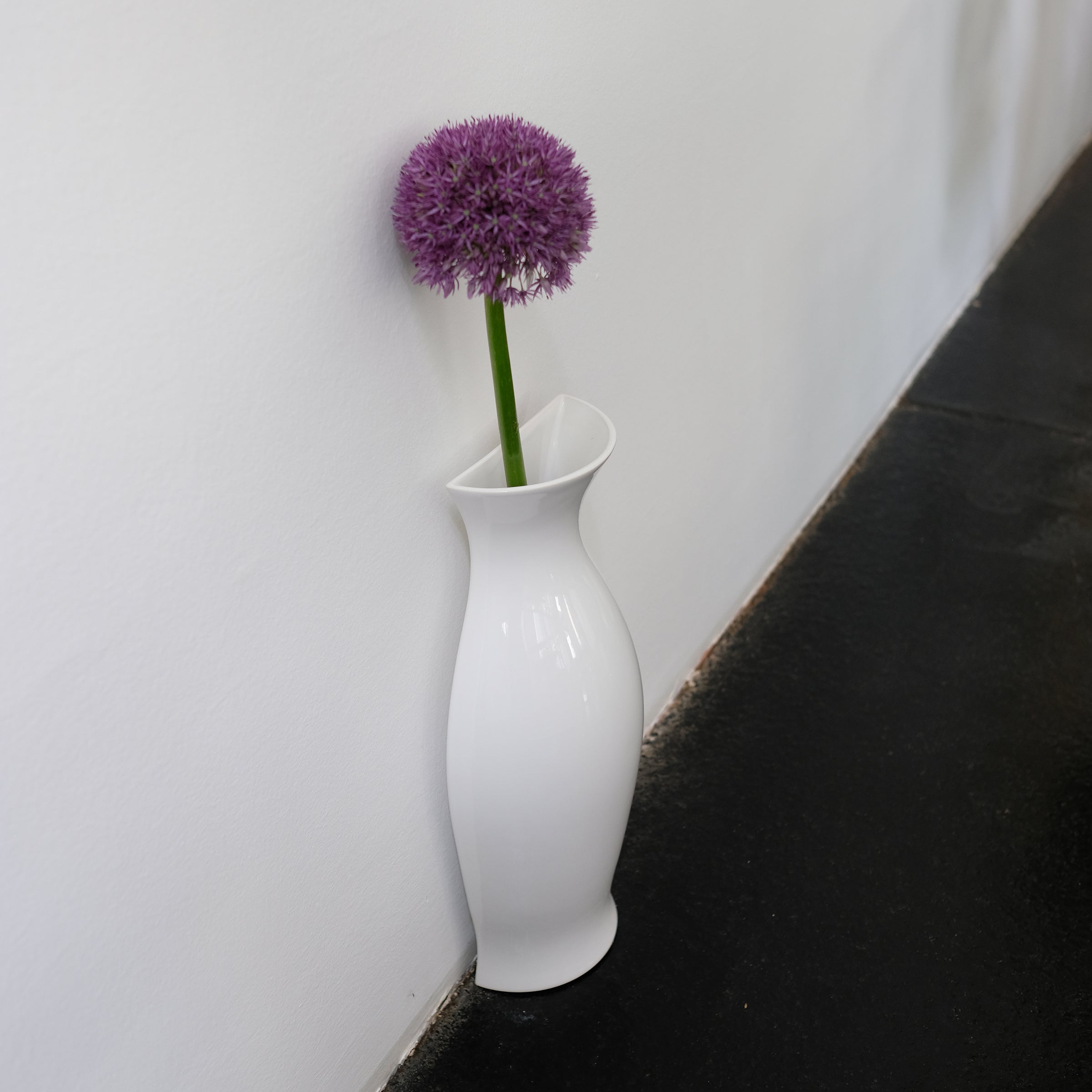 White glossy ceramic QUATER Vase with a single purple flower, standing against a white wall on a dark floor.