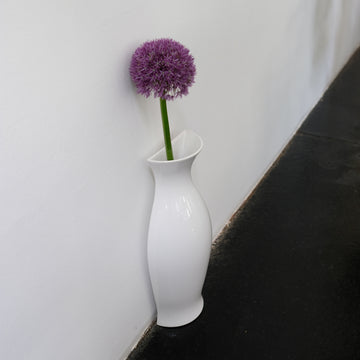 White glossy ceramic QUATER Vase with a single purple flower, standing against a white wall on a dark floor.