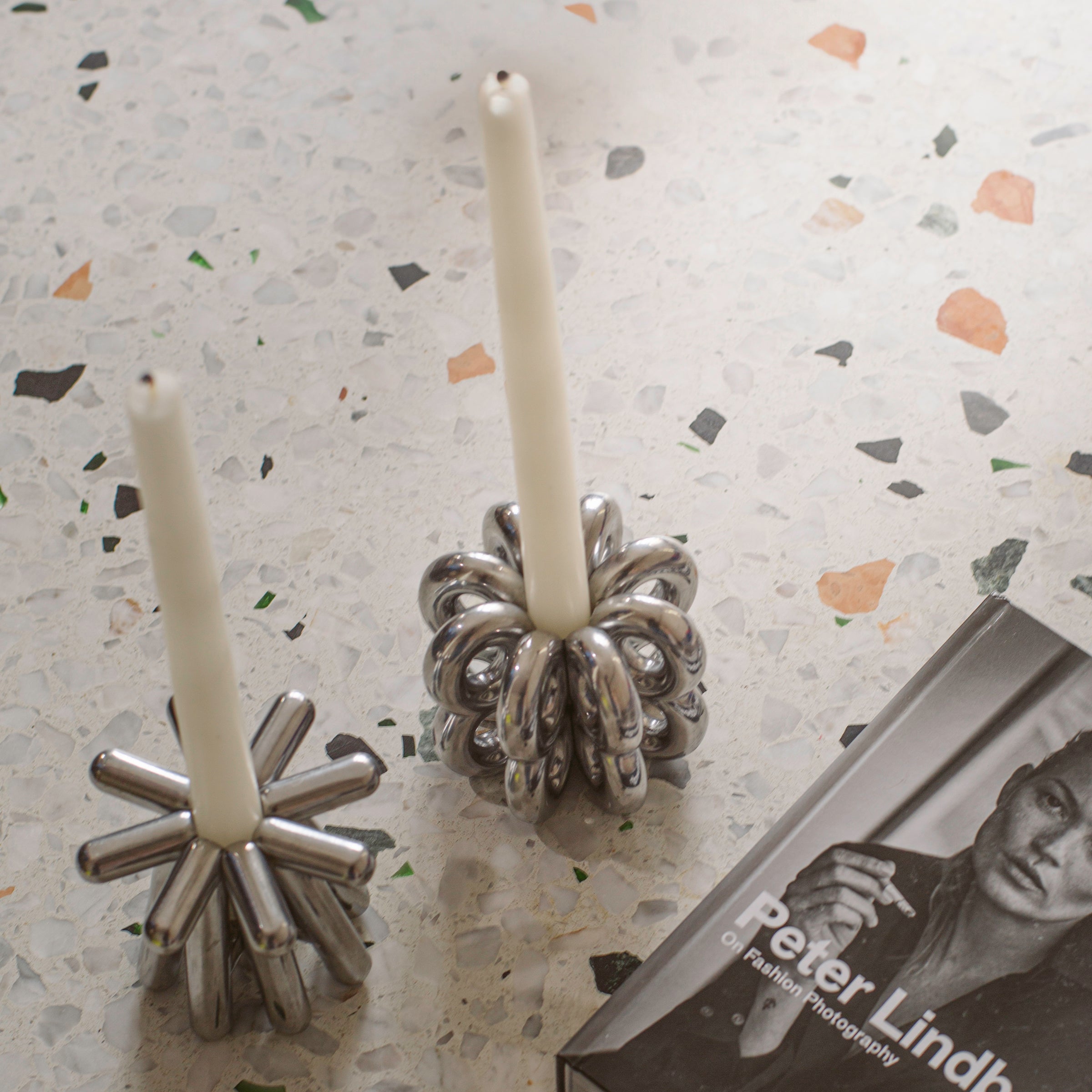 Close-up of two silver DIGIT NINE Candlestick holders with white candles on a terrazzo surface next to a book.