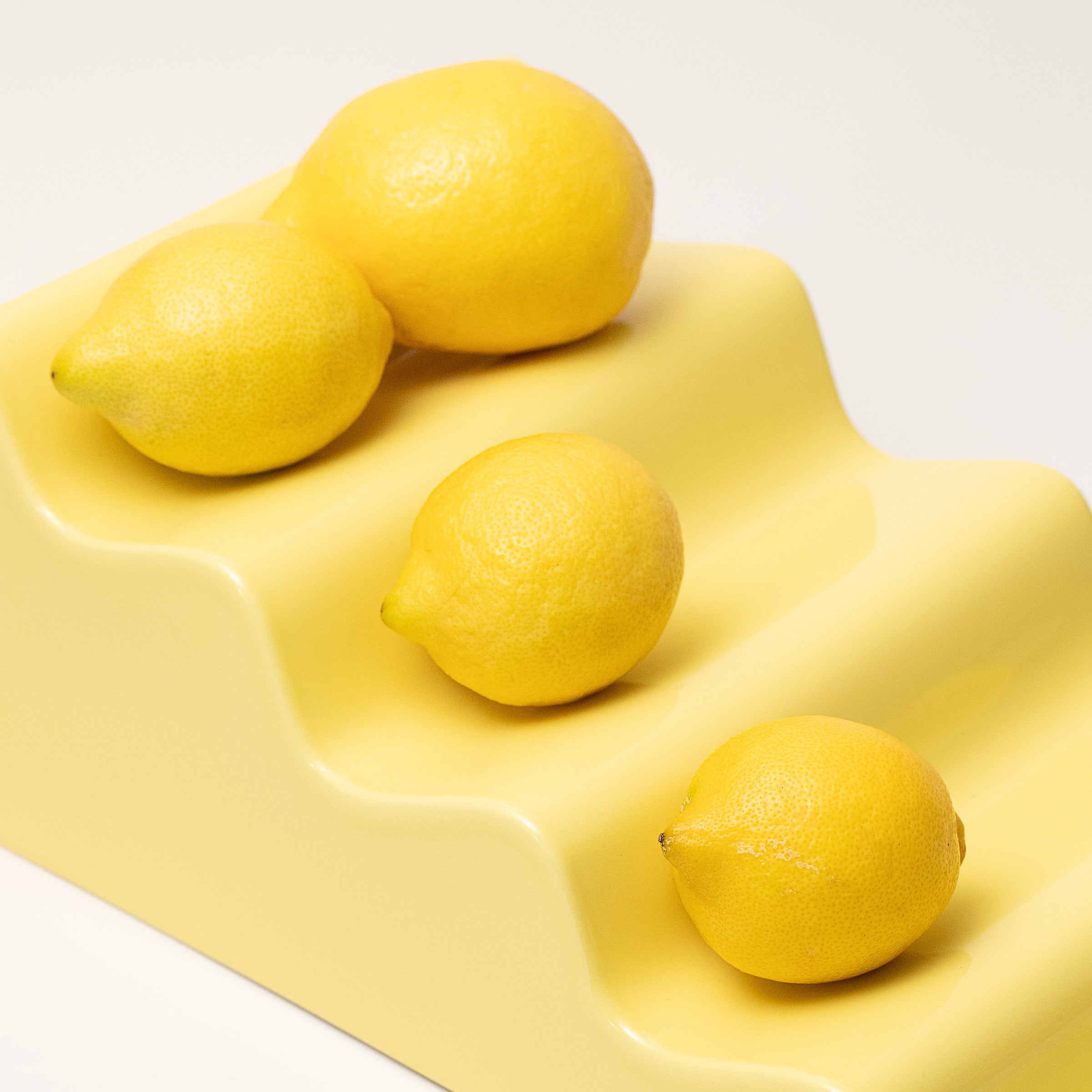 CASCADE Fruitstand in glossy yellow ceramic holding four fresh lemons in its wavy slots.