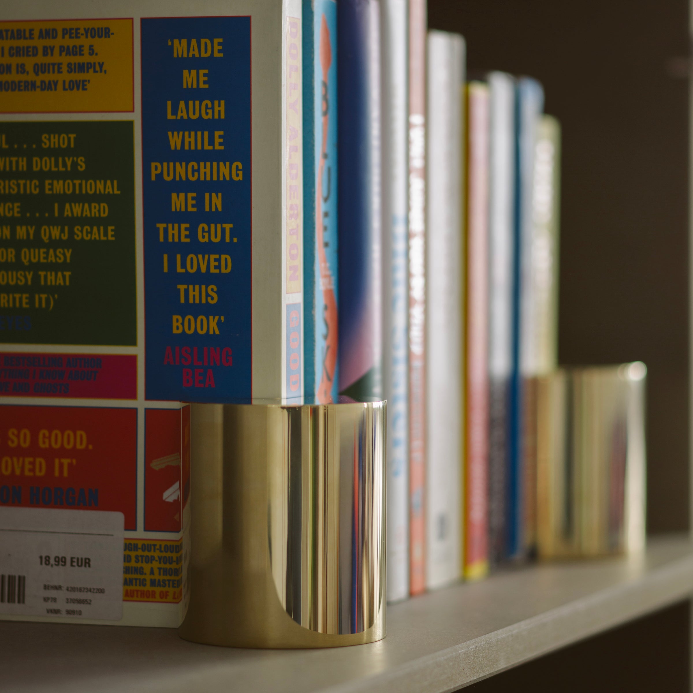 NOOK Bookend A polished brass bookend holding upright a row of mixed colorful books on a shelf.