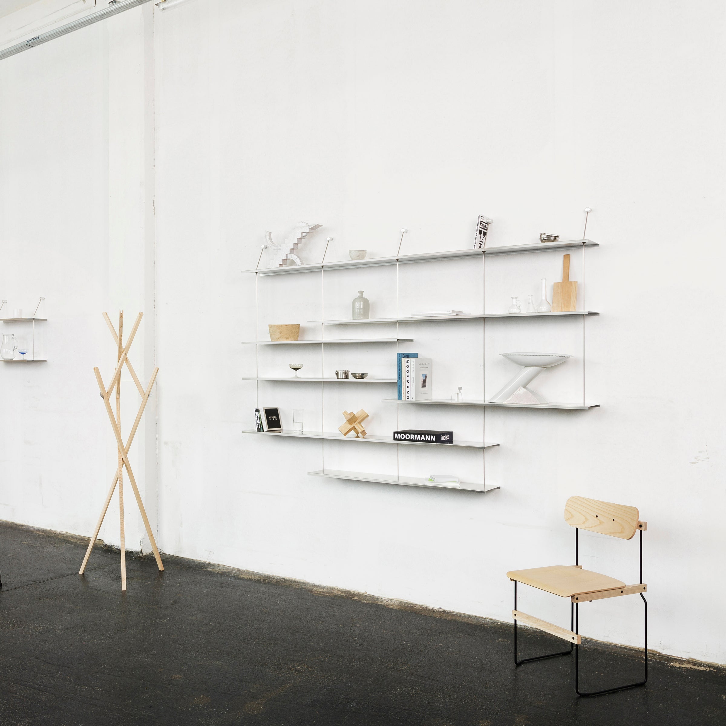 A minimalist room features Liesl by Moormann, a shelf combination displaying decor and books on a white wall. A simple wooden coat rack and chair with metal legs complements the space, all set on a dark floor.