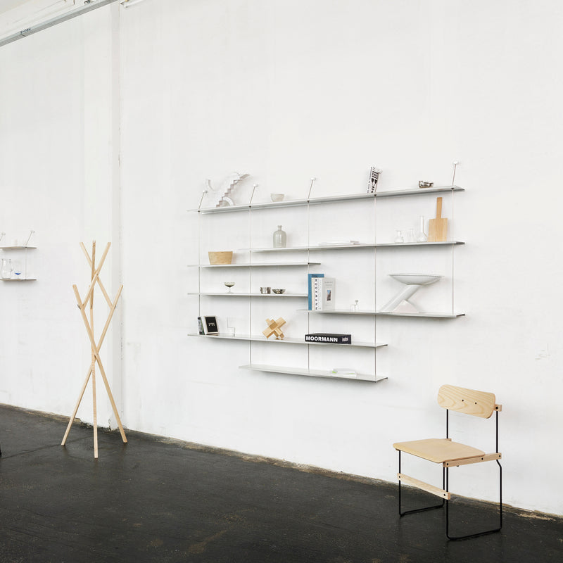 A minimalist room features Liesl by Moormann, a shelf combination displaying decor and books on a white wall. A simple wooden coat rack and chair with metal legs complements the space, all set on a dark floor.