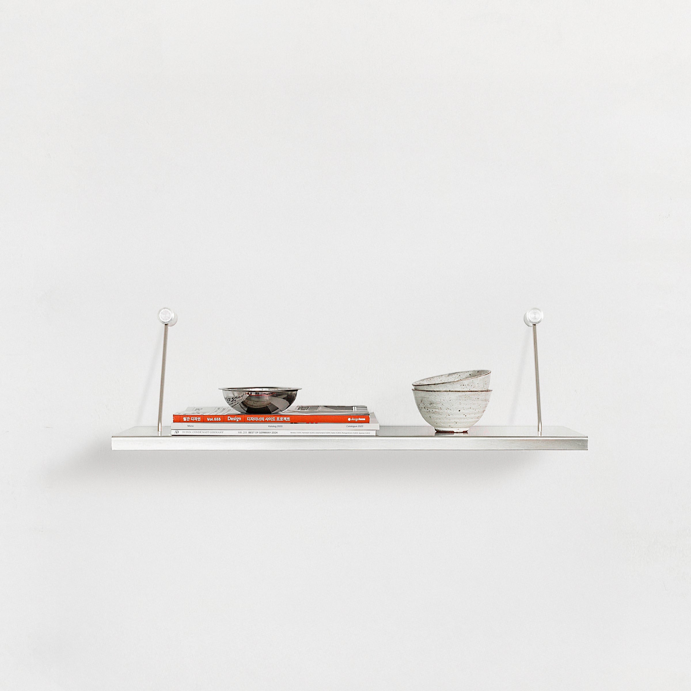 The Moormann Liesl shelf, mounted on a white wall, displays two stacked books topped with a metallic bowl and features two ceramic bowls, one speckled and tilted, creating a simple and modern look.