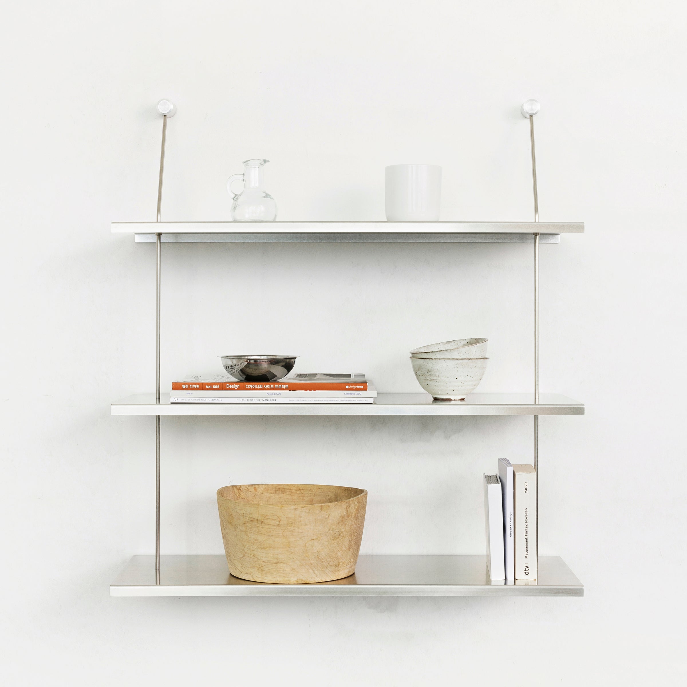 The Liesl by Moormann is a minimalist, wall-mounted, three-tier shelf. The top shelf holds a glass jar and white mug; the middle has a silver bowl, books, and a ceramic bowl; and the bottom shows off a wooden bowl and upright books against a crisp white background—ideal for modular furniture fans.