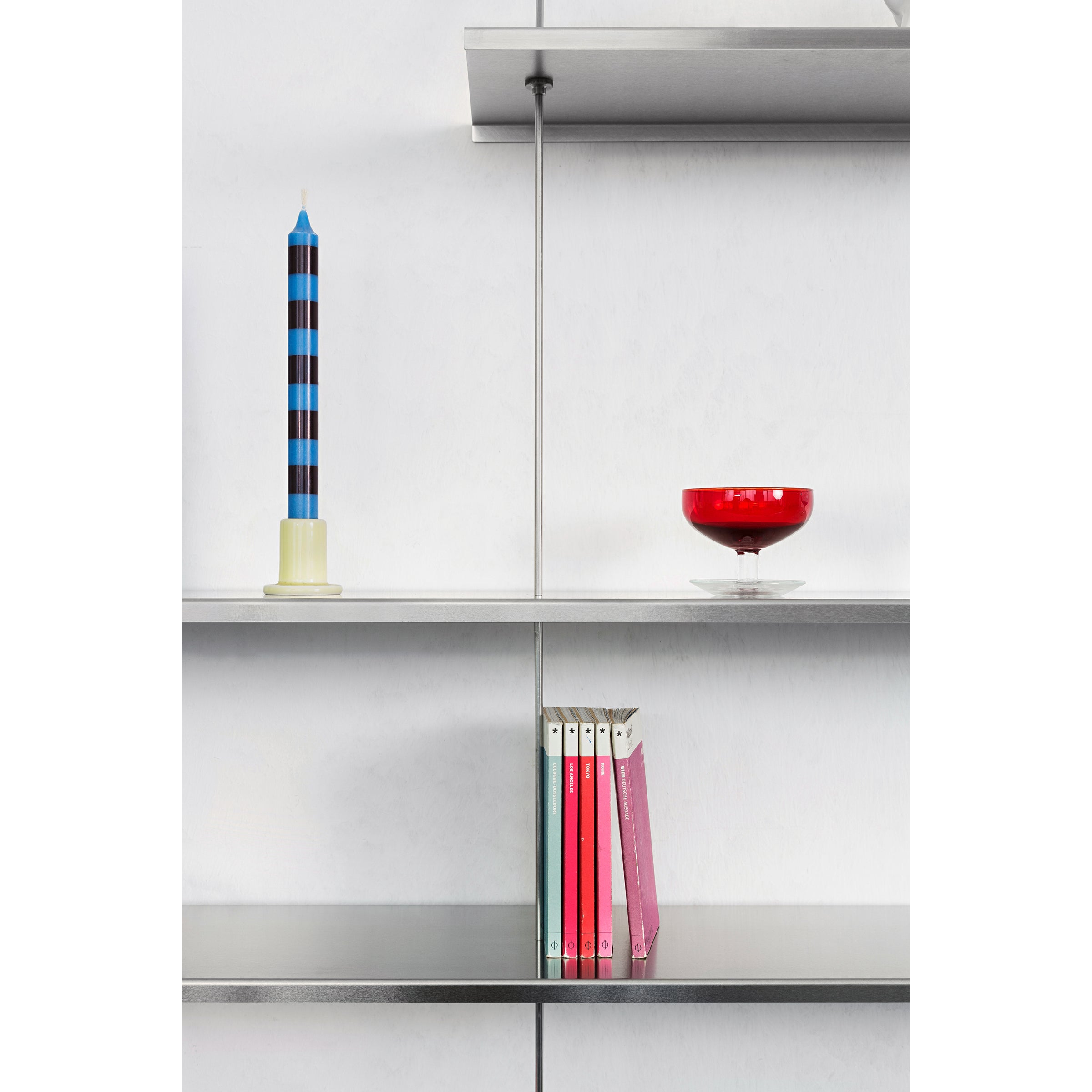 The Moormann Liesl, a modern wall-mounted modular shelving unit, showcases a tall striped candle on the top shelf, a red glass bowl in the middle, and five books with colorful spines on the bottom shelf against a plain white wall.