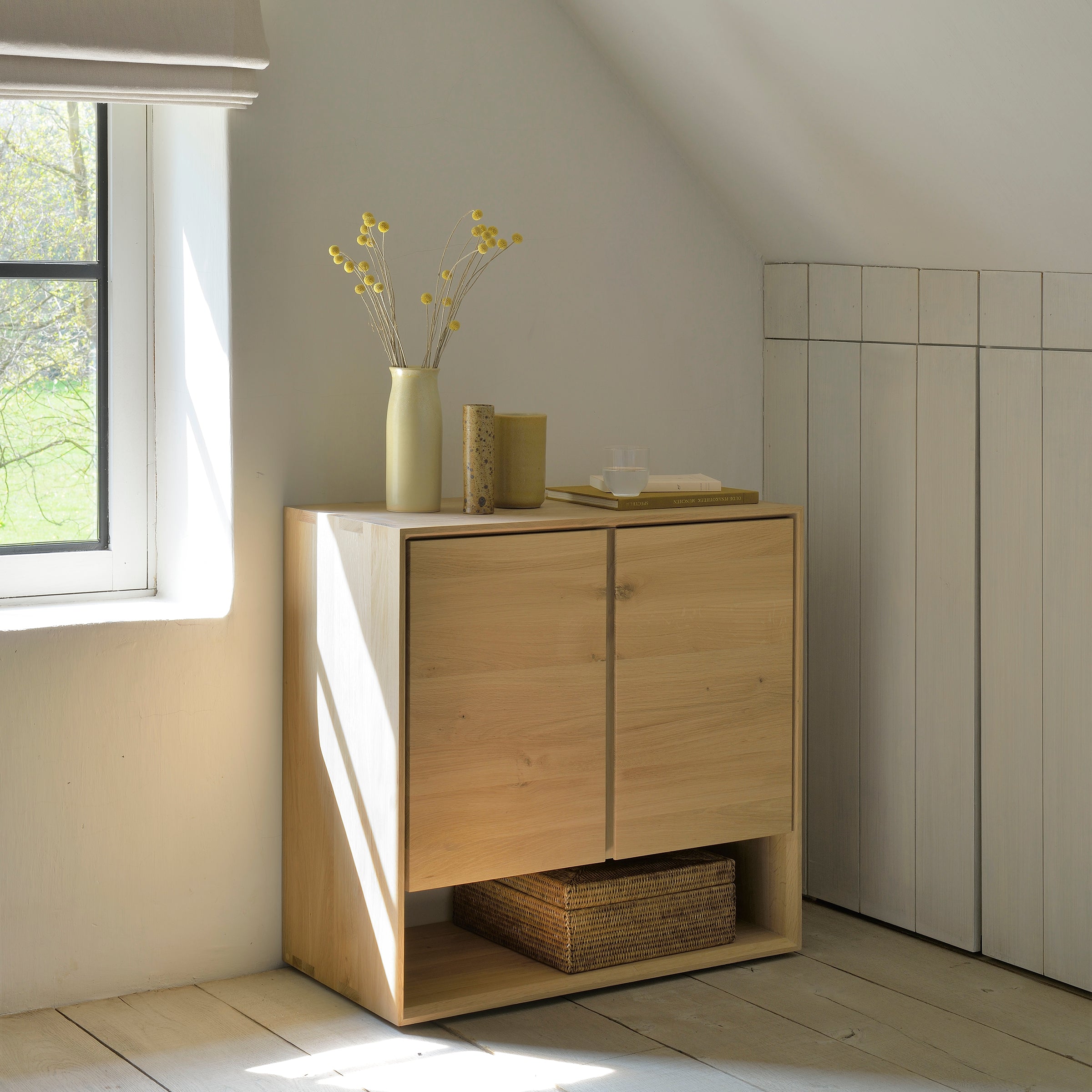 The Ethnicraft Nordic Sideboard stands by a sunlit window, topped with a vase of yellow flowers, books, and glasses. A woven basket rests below—all in a minimalist room, its beveled edges adding subtle detail.