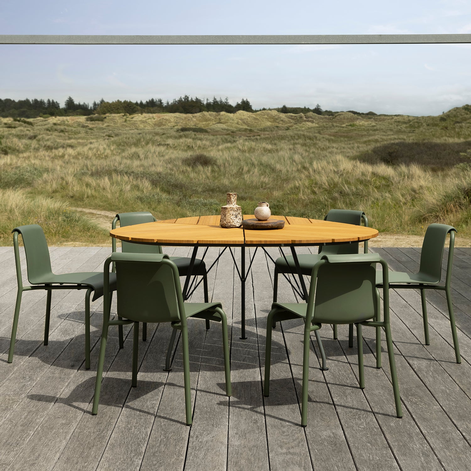 A round wooden table with a decorative vase and bowl is surrounded by six Nami Dining Chairs without armrests by Houe on a gray wooden deck. The elegant powder-coated steel frames complement the setting, with grassy dunes and a forest under a cloudy sky as a serene backdrop.