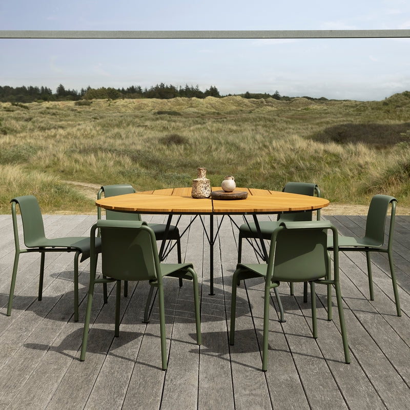 A round wooden table with a decorative vase and bowl is surrounded by six Nami Dining Chairs without armrests by Houe on a gray wooden deck. The elegant powder-coated steel frames complement the setting, with grassy dunes and a forest under a cloudy sky as a serene backdrop.
