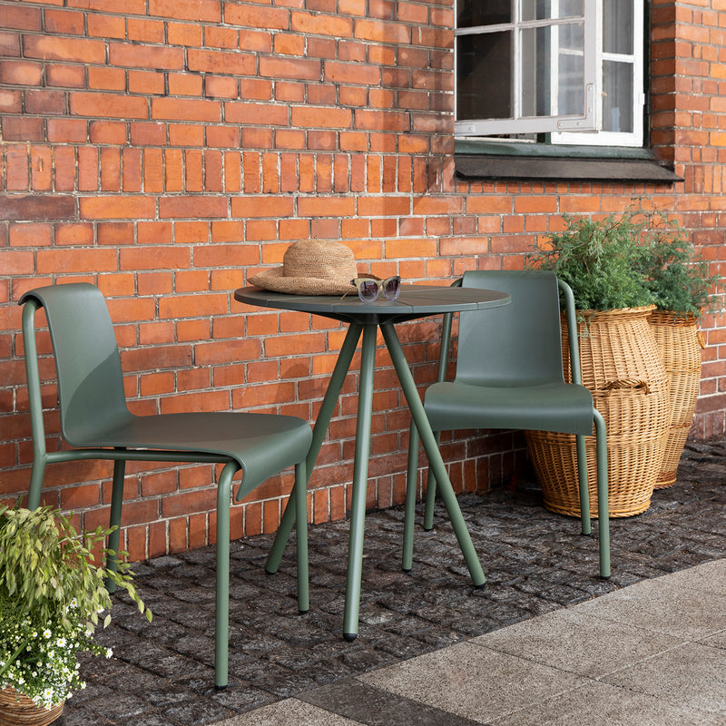 Two Nami Dining Chairs by Houe, in green, and a round powder-coated steel table are placed on a stone patio against a brick wall. A hat and sunglasses lie on the table. A wicker basket filled with plants adds greenery, with a window gleaming above this charming arrangement.