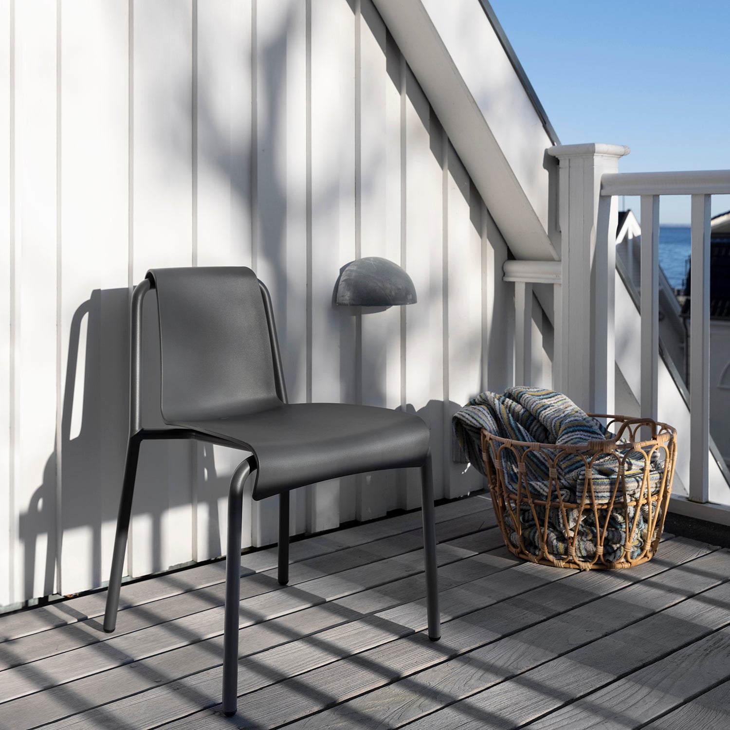 A Nami Dining Chair without Armrest by Houe and a woven basket with striped cushions rest on a sunlit wooden deck. The deck, featuring powder-coated steel railings, is surrounded by white panels, against a backdrop of clear blue sky.