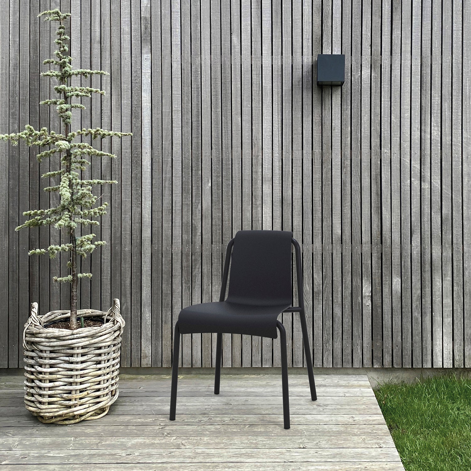 A black Nami Dining Chair without Armrest by Houe, crafted from powder-coated steel, sits elegantly on a wooden deck next to a tree in a basket-style planter. Vertical wood planks serve as the backdrop, with a small black wall light fixture above.