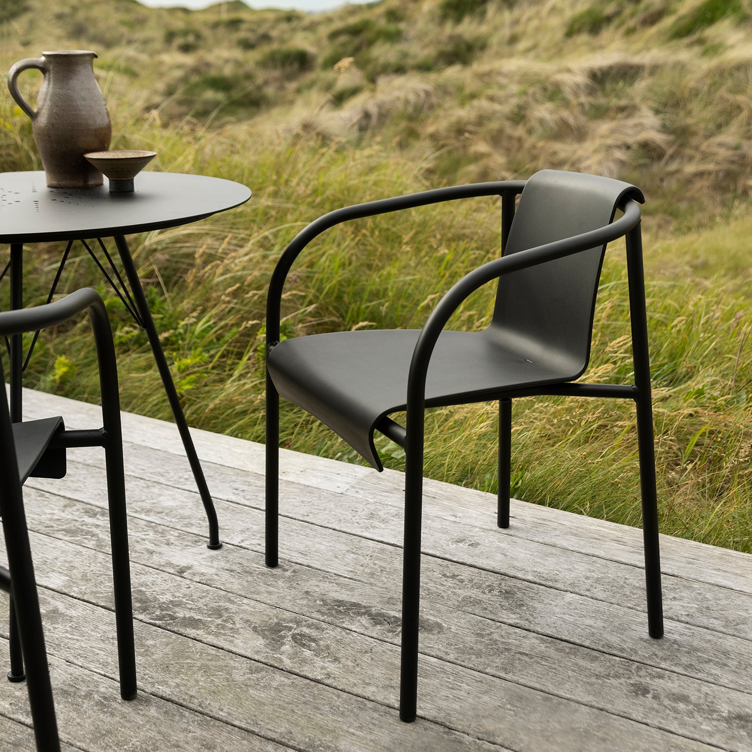 The Houe Nami Dining Chair with Armrest in powder-coated steel pairs beautifully with a round table featuring a ceramic jug and bowl, creating a chic setup on the wooden patio. In the background, tall grass sways gently against rolling hills under a cloudy sky.