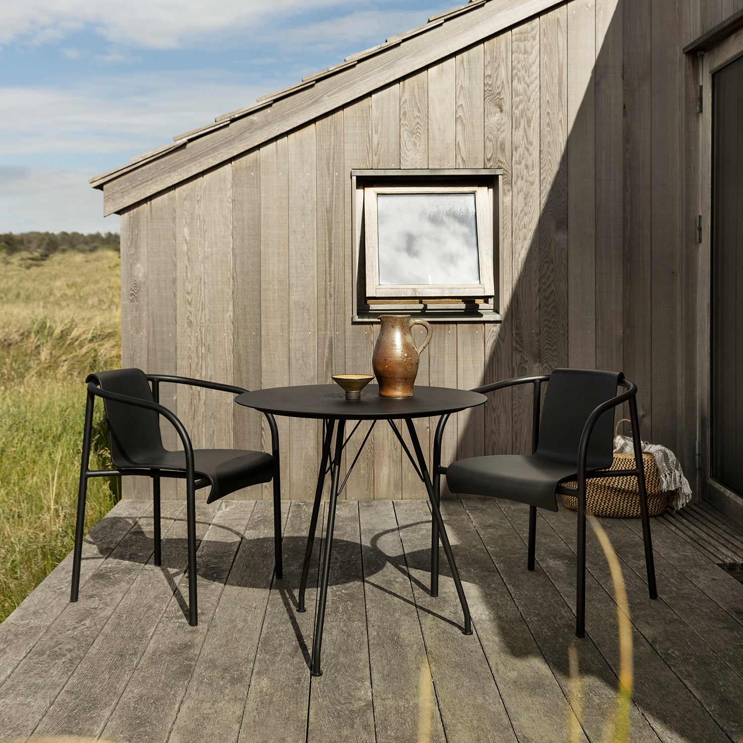 A small outdoor patio set with a round powder-coated steel table and two matching Nami Dining Chairs with Armrest by Houe sits on a wooden deck. The table holds a pitcher and bowl, while a wooden building overlooks grassy fields under a blue sky.