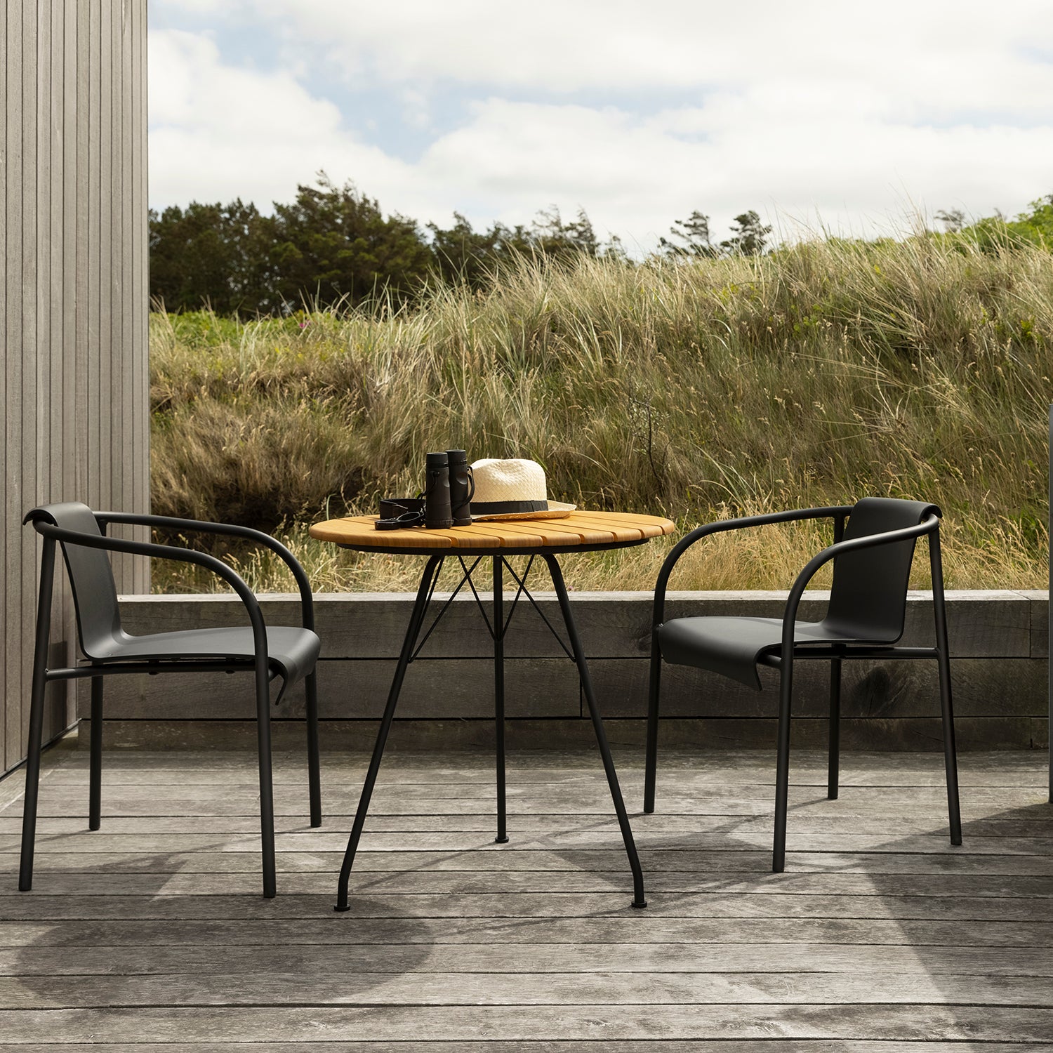 On a deck sits a wooden outdoor table with two Houe Nami Dining Chairs. A camera, hat, and cup are on the table as a grassy field extends into the cloudy skys backdrop, while the powder-coated steel frames of the chairs add modern elegance.