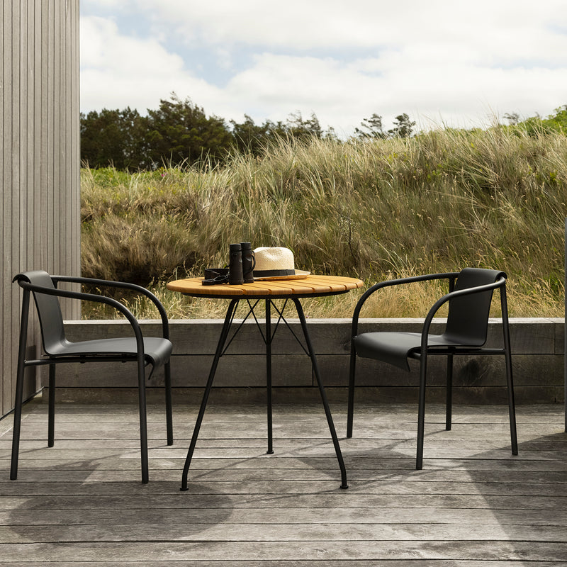 On a deck sits a wooden outdoor table with two Houe Nami Dining Chairs. A camera, hat, and cup are on the table as a grassy field extends into the cloudy skys backdrop, while the powder-coated steel frames of the chairs add modern elegance.