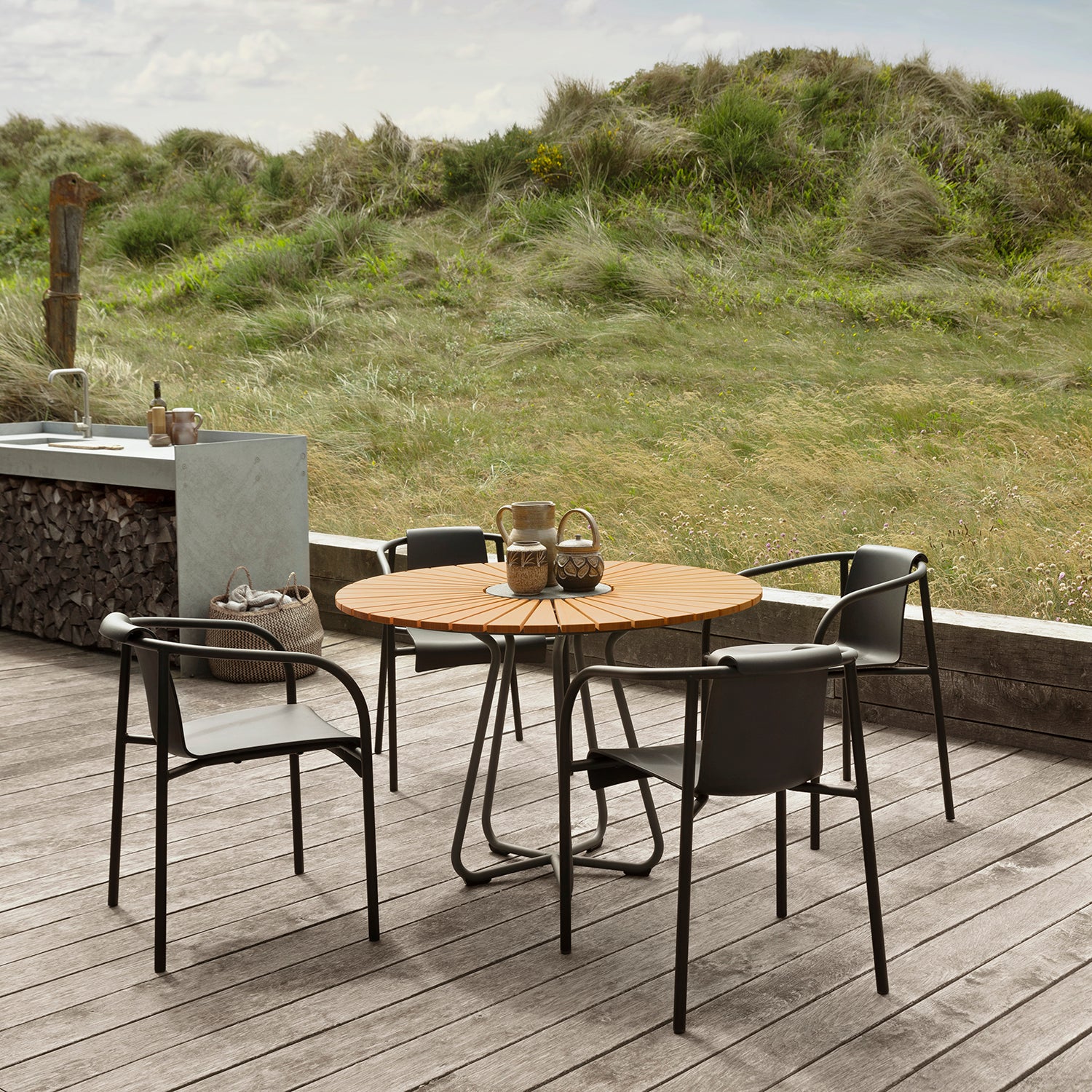 A sunlit wooden deck features a circular table paired with Houes Nami Dining Chairs with Armrests. The scene is set against a grassy landscape, stone counter adorned with a basket and firewood stack nearby, all under a partly cloudy sky complemented by garden chairs.
