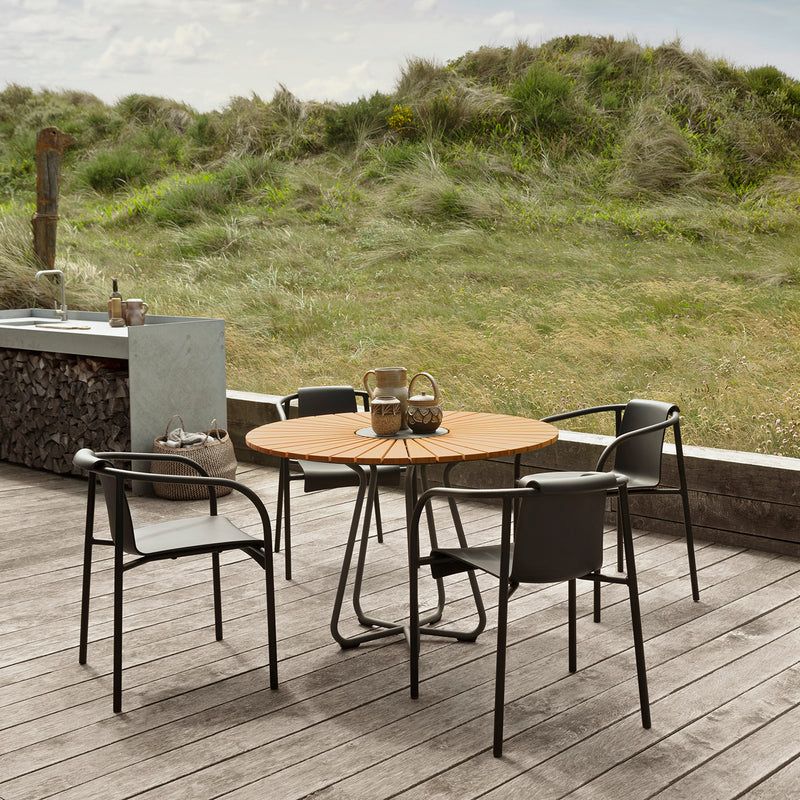 A sunlit wooden deck features a circular table paired with Houes Nami Dining Chairs with Armrests. The scene is set against a grassy landscape, stone counter adorned with a basket and firewood stack nearby, all under a partly cloudy sky complemented by garden chairs.