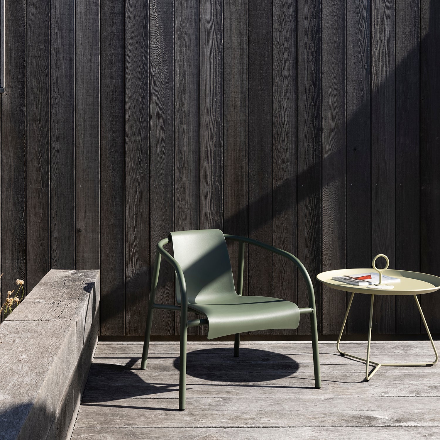 A modern outdoor setup includes a Nami Lounge Chair by Houe, made from Danish plastic waste and powder-coated steel, alongside a magazine-laden round table on the wooden deck. A dark wooden wall and stone bench with shifting shadows complete the minimalist scene.