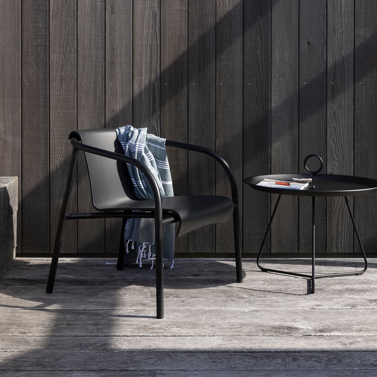 A modern black Nami Lounge Chair by Houe, crafted from Danish plastic waste, is draped with a blue and white blanket. Beside it, a round powder-coated steel table supports an open magazine. Placed against a wooden wall, the outdoor setting enjoys sunlight that casts playful shadows on the ground.