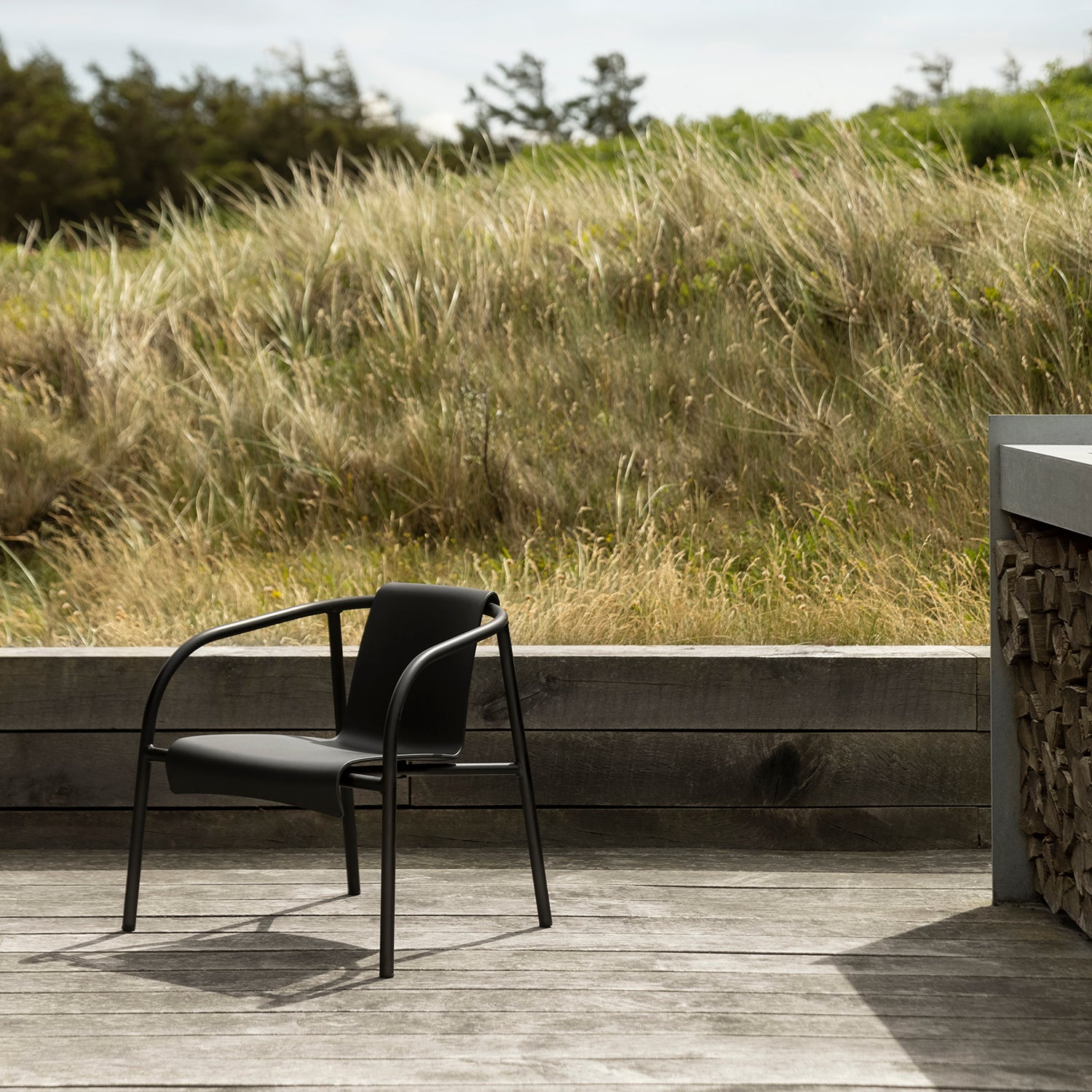 A modern black Nami Lounge Chair by Houe is positioned on a wooden deck, facing grassy dunes. A stack of firewood is partly visible to the right near a concrete wall, and trees in the background are under a cloudy sky.