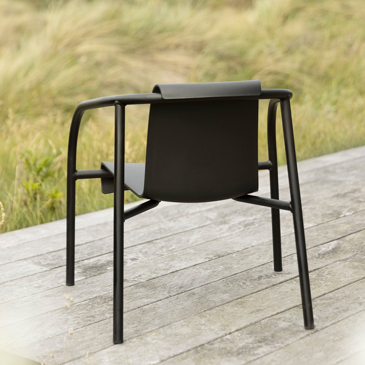 The Nami Lounge Chair by Houe, featuring a curved frame and flat seat in contemporary style, crafted from powder-coated steel, sits on a wooden deck. The deck is set against blurred tall grasses, enhancing the serene natural setting.