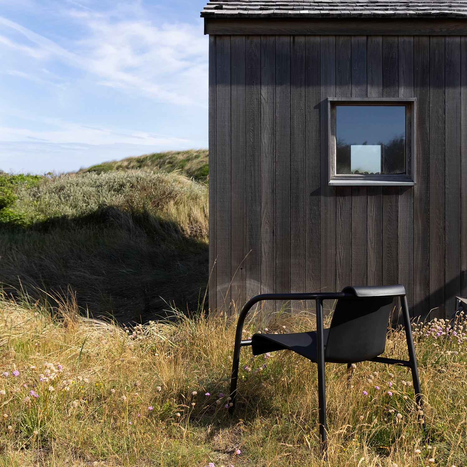 A single Nami Lounge Chair by Houe, crafted from Danish plastic waste, rests on grass near a wooden building with a small window, set against lush hills and a partly cloudy sky.